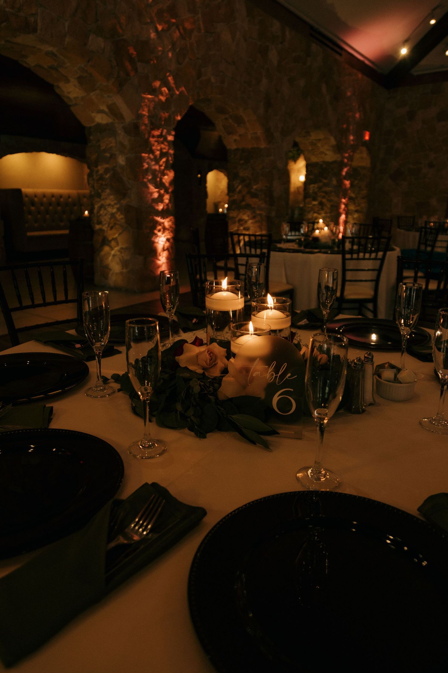 Romantic candlelit wedding reception tables with black plates, floral centerpieces in rustic stone venue.