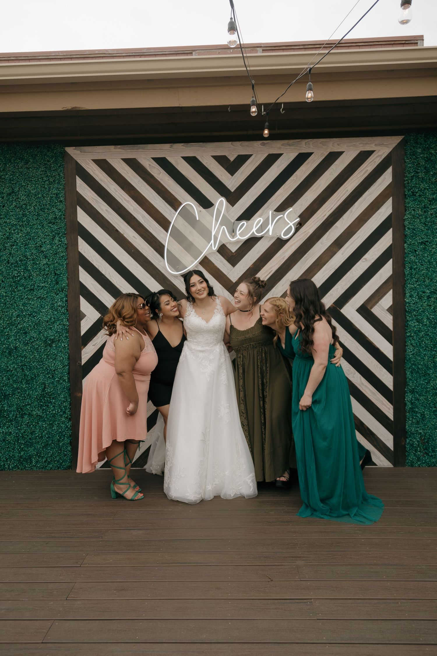 Bride poses with bridesmaids in colorful gowns in front of a Cheers neon sign backdrop.