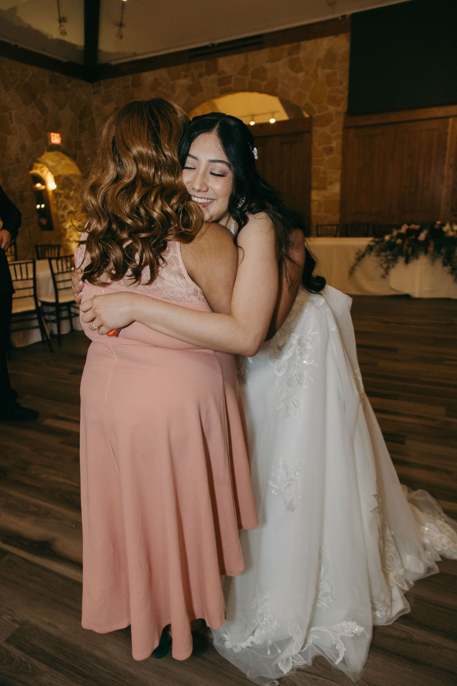 A smiling bride in a white lace gown shares a warm embrace with a guest in a pink dress.