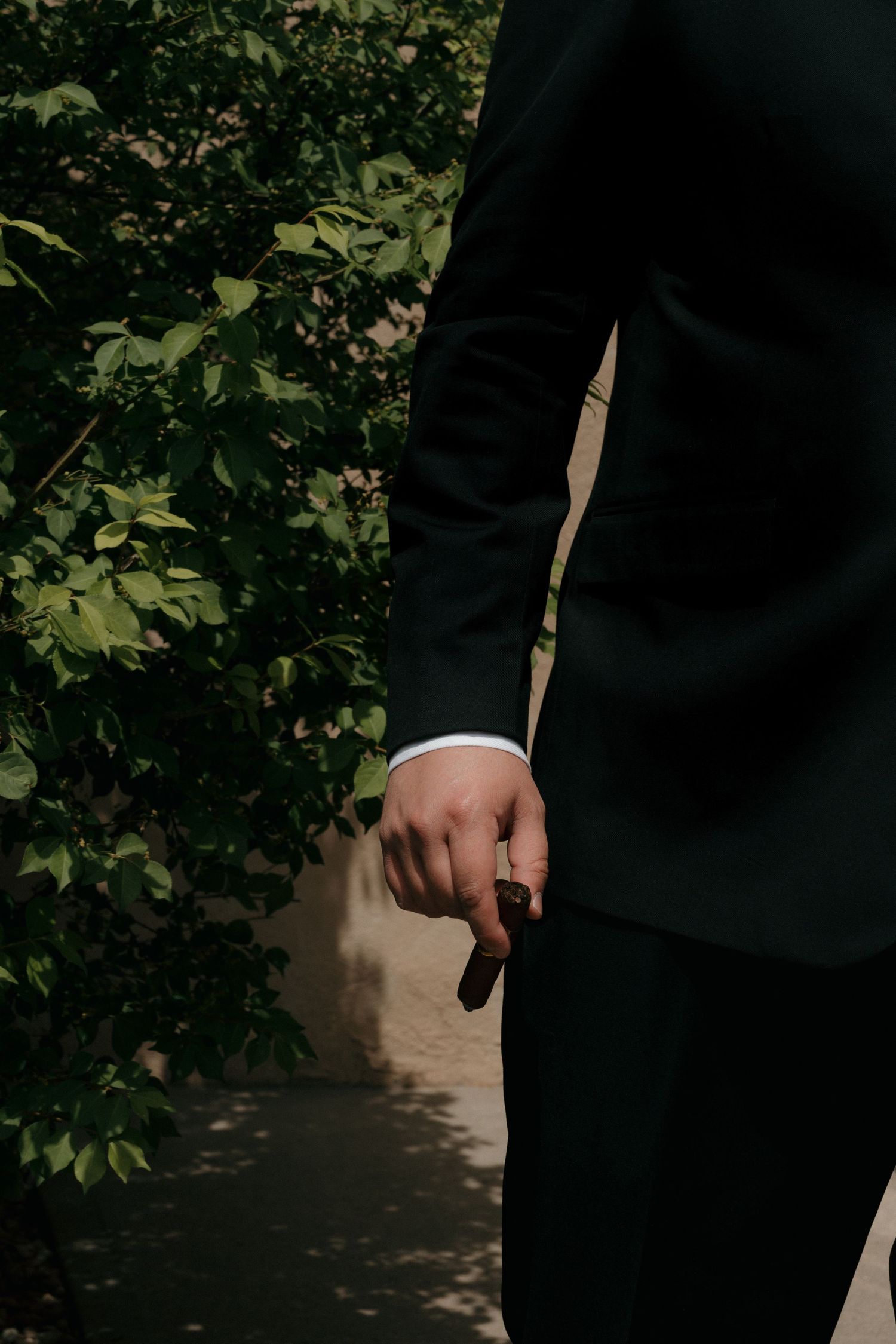 Groom in dark green suit holding a ring outdoors near lush green foliage in natural sunlight.