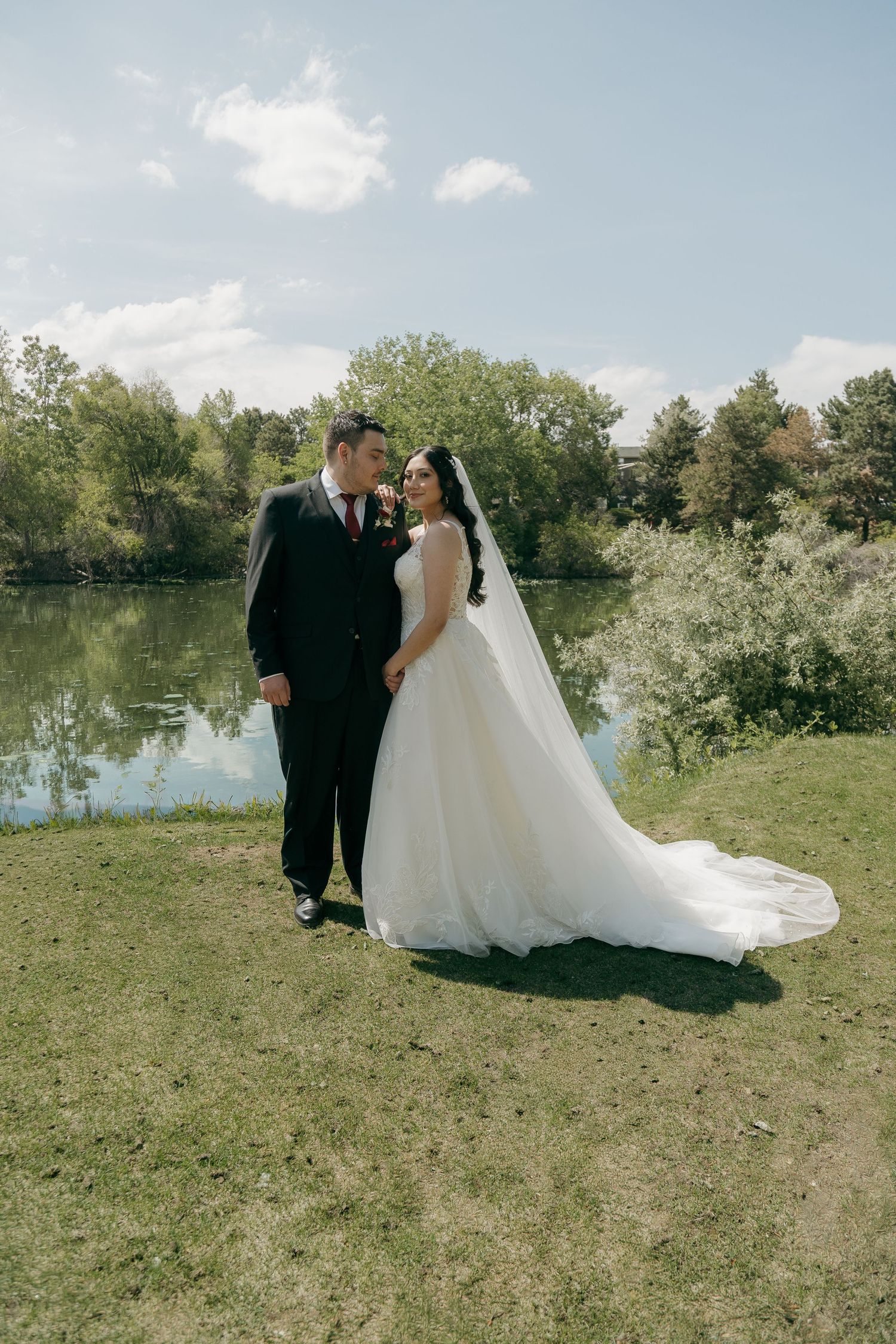 Bride and groom pose romantically by a serene lake surrounded by lush green trees on their wedding day.