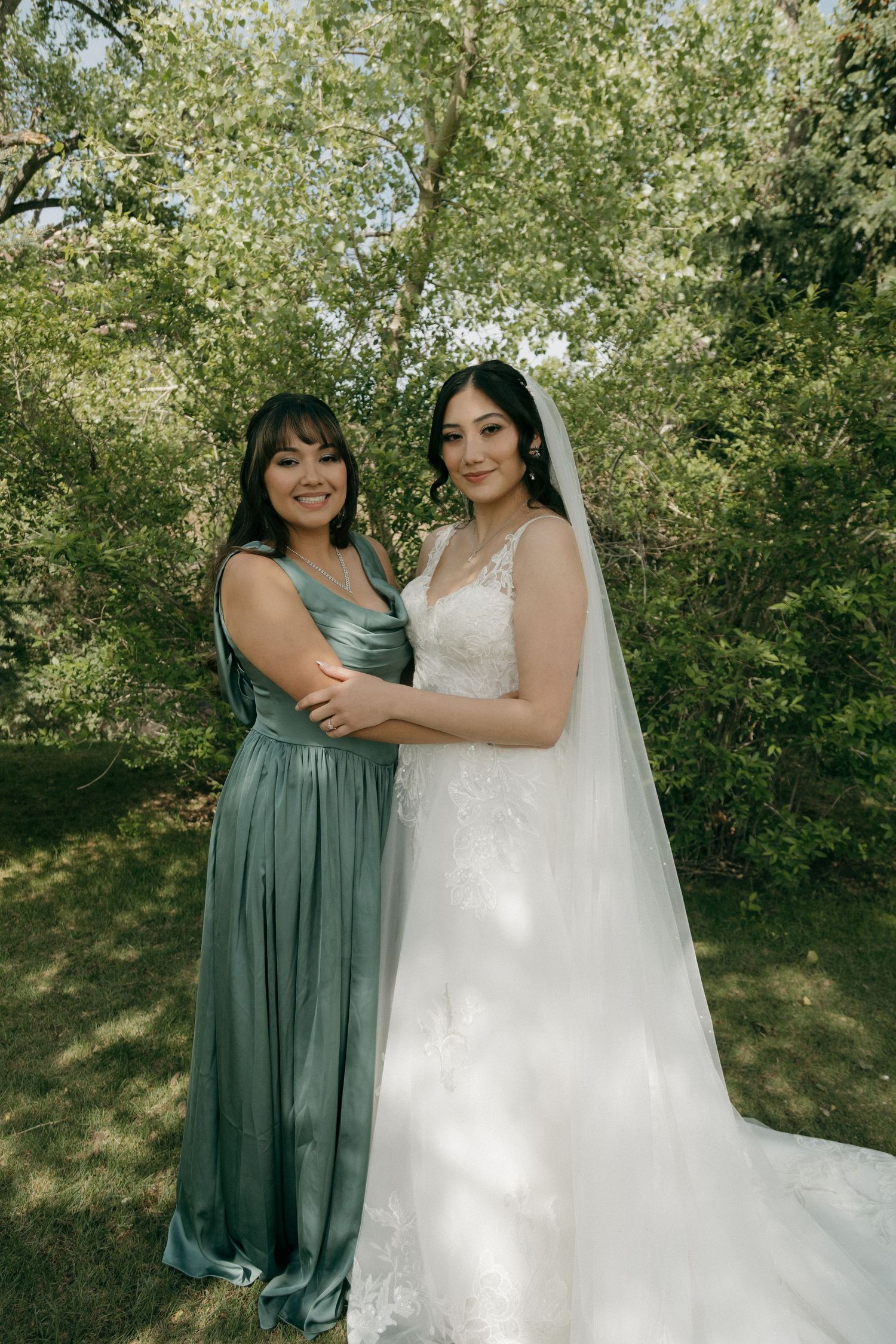 Bride in white lace gown with veil poses with bridesmaid in sage green dress outdoors.
