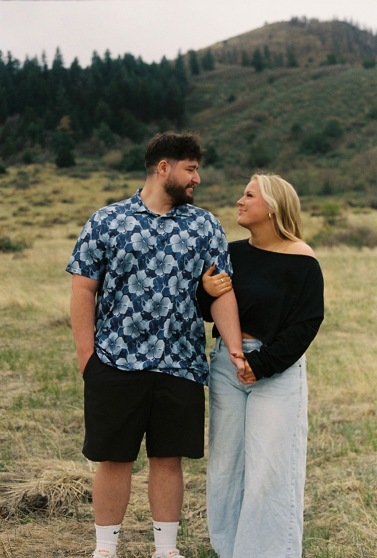 Couple holding hands and smiling at each other in a grassy meadow with green hills and pine trees in the background.