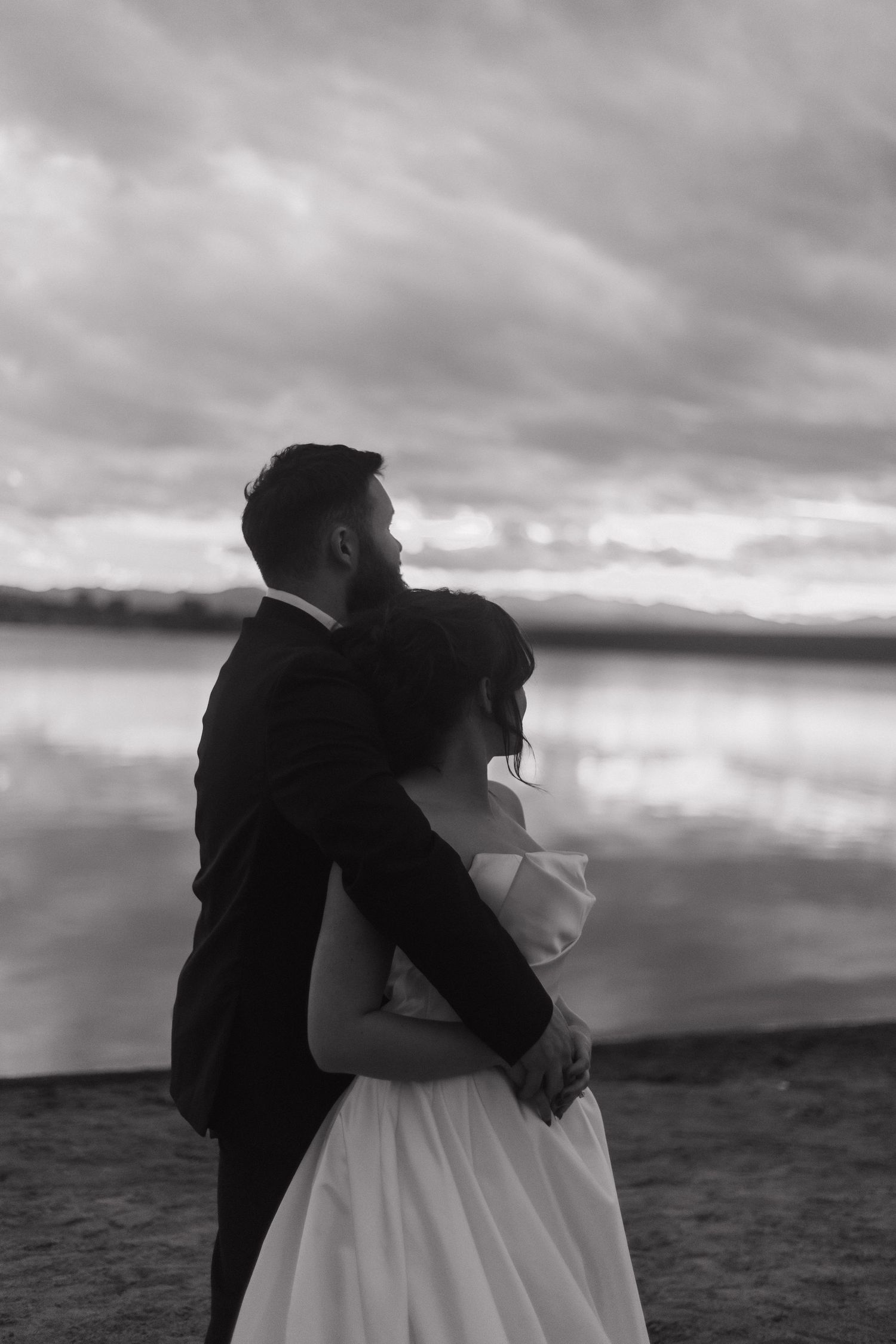 A romantic black and white portrait of a couple embracing on a cloudy beach.