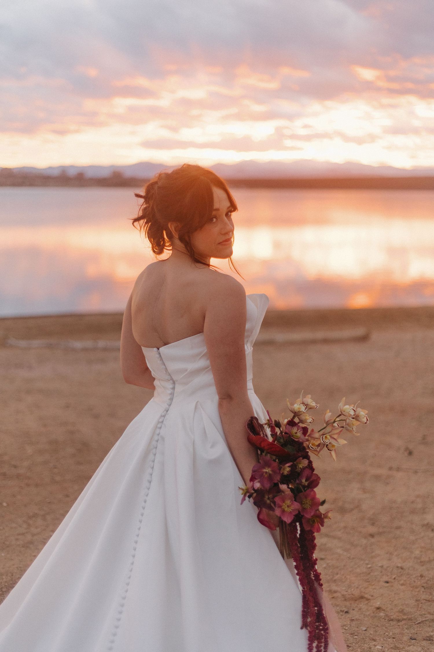 Bride in a white wedding dress holding a bouquet against a sunset beach backdrop.