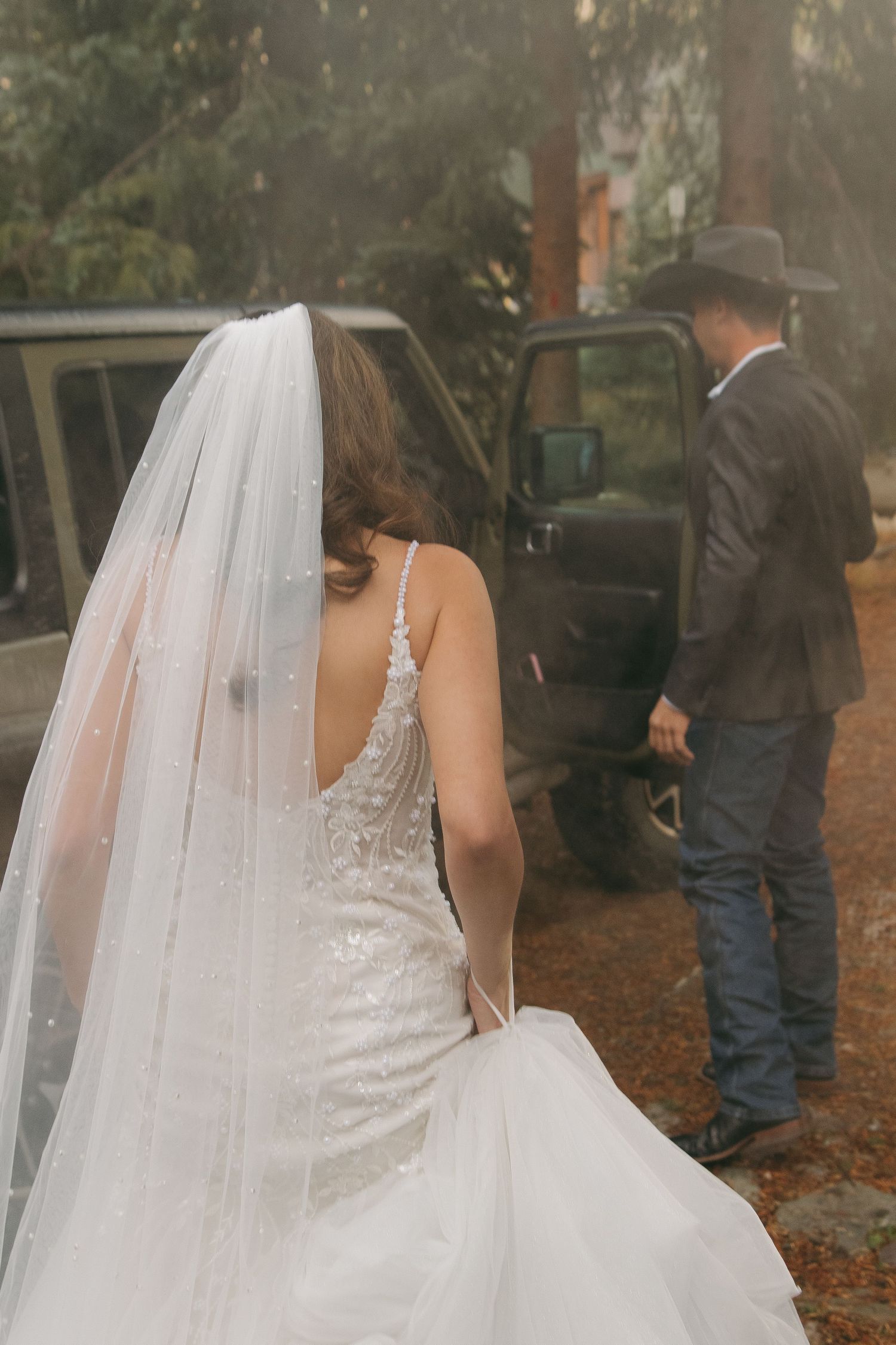 A bride in a lace wedding dress and veil stands near a vehicle in a wooded setting.