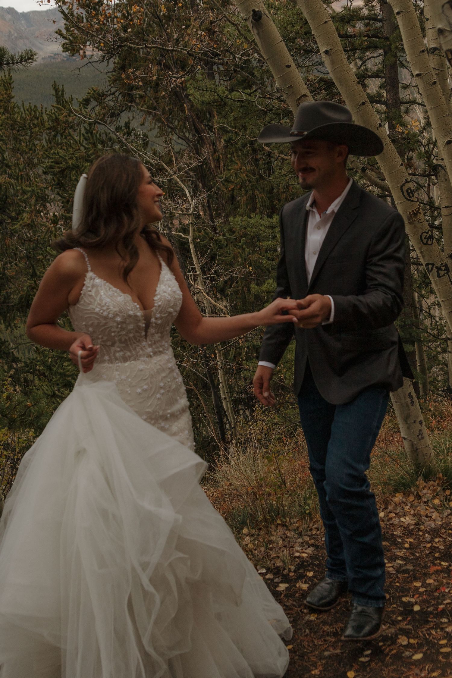 A couple dancing playfully in a wooded area with autumn foliage, the bride wearing a flowing white gown.
