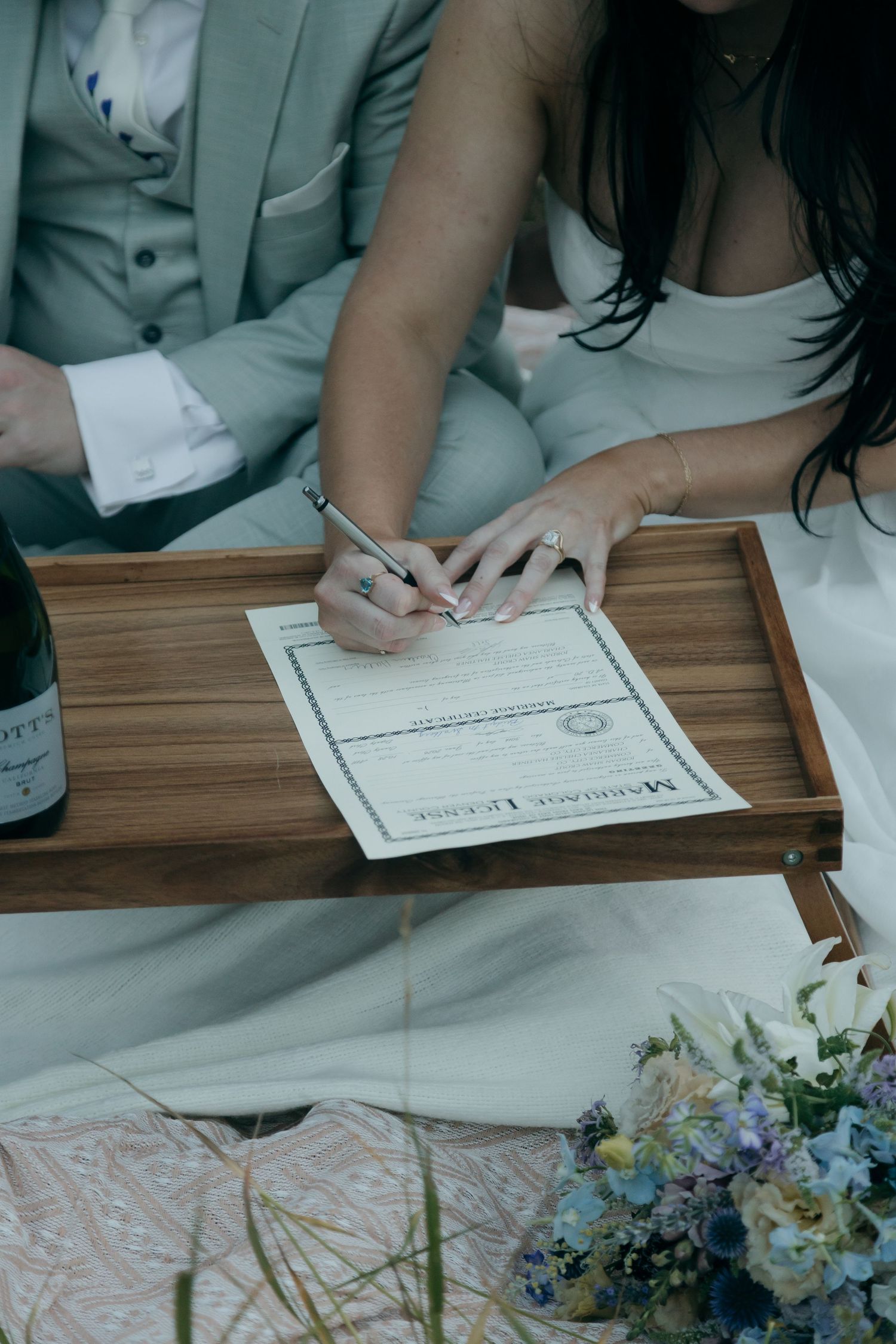 Bride signing marriage license on wooden tray during outdoor elopement ceremony with champagne and wildflower bouquet.
