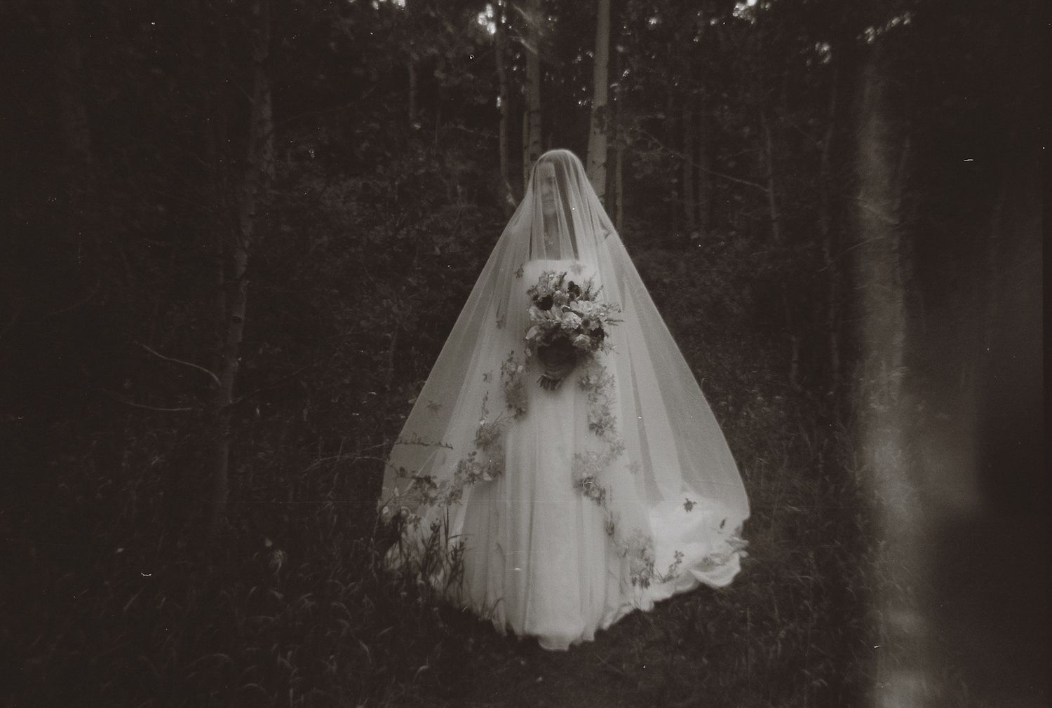 Ethereal black and white film photo of a veiled bride holding a bouquet standing alone in a dark forest.