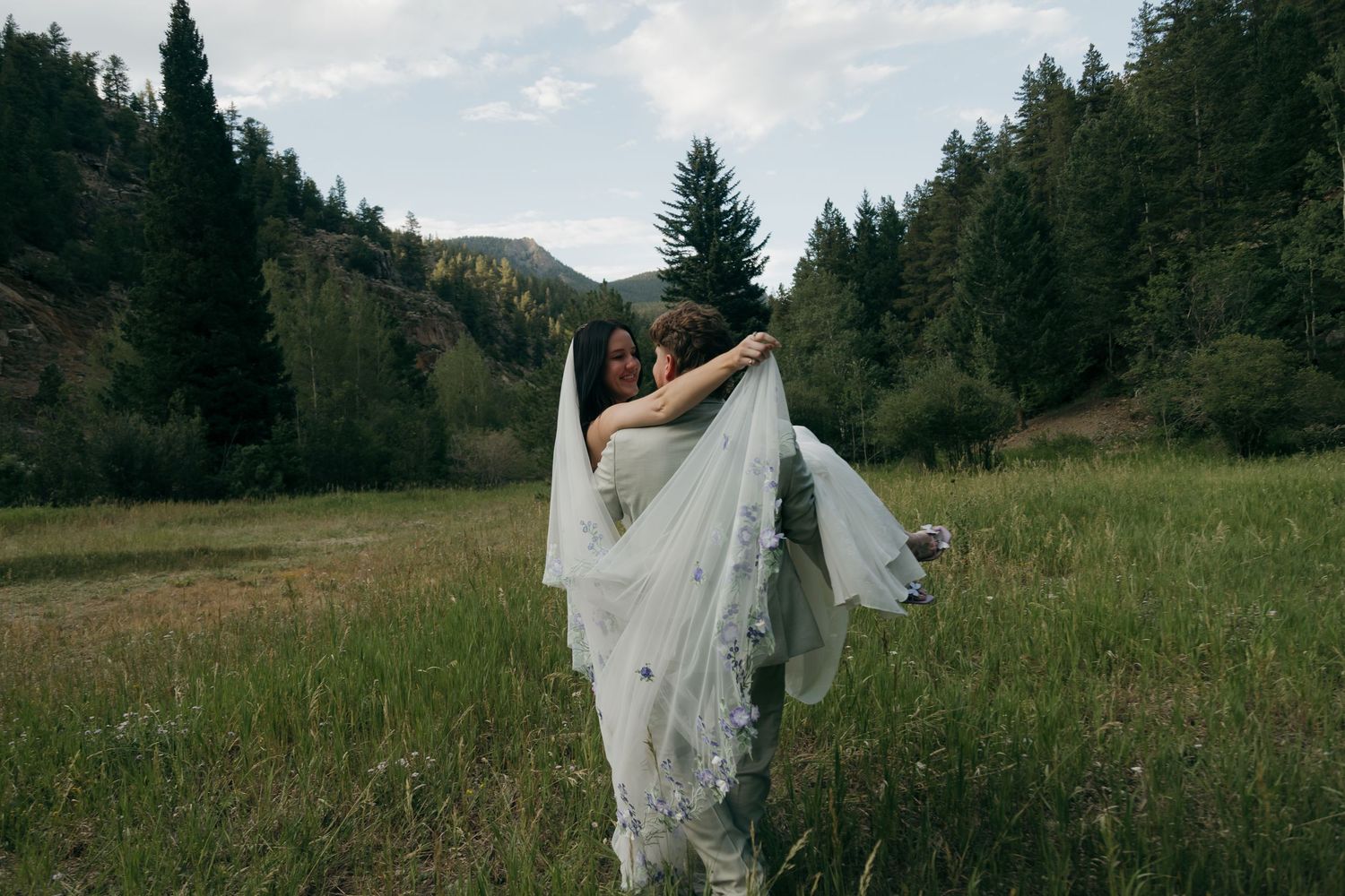 Bride and groom share a romantic embrace in a green mountain meadow, with the bride's white veil flowing in the breeze.