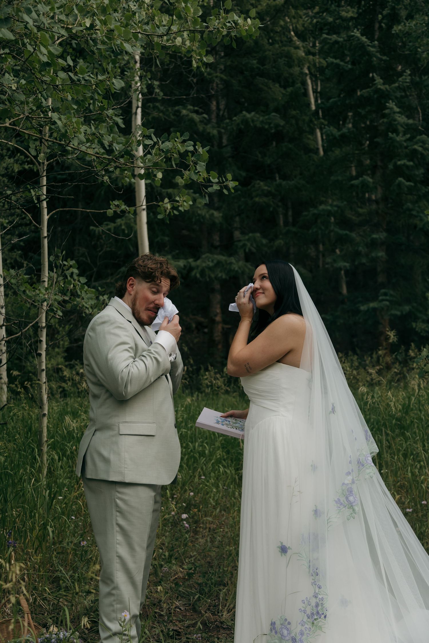 Emotional bride and groom wipe tears during outdoor forest wedding ceremony vow exchange.