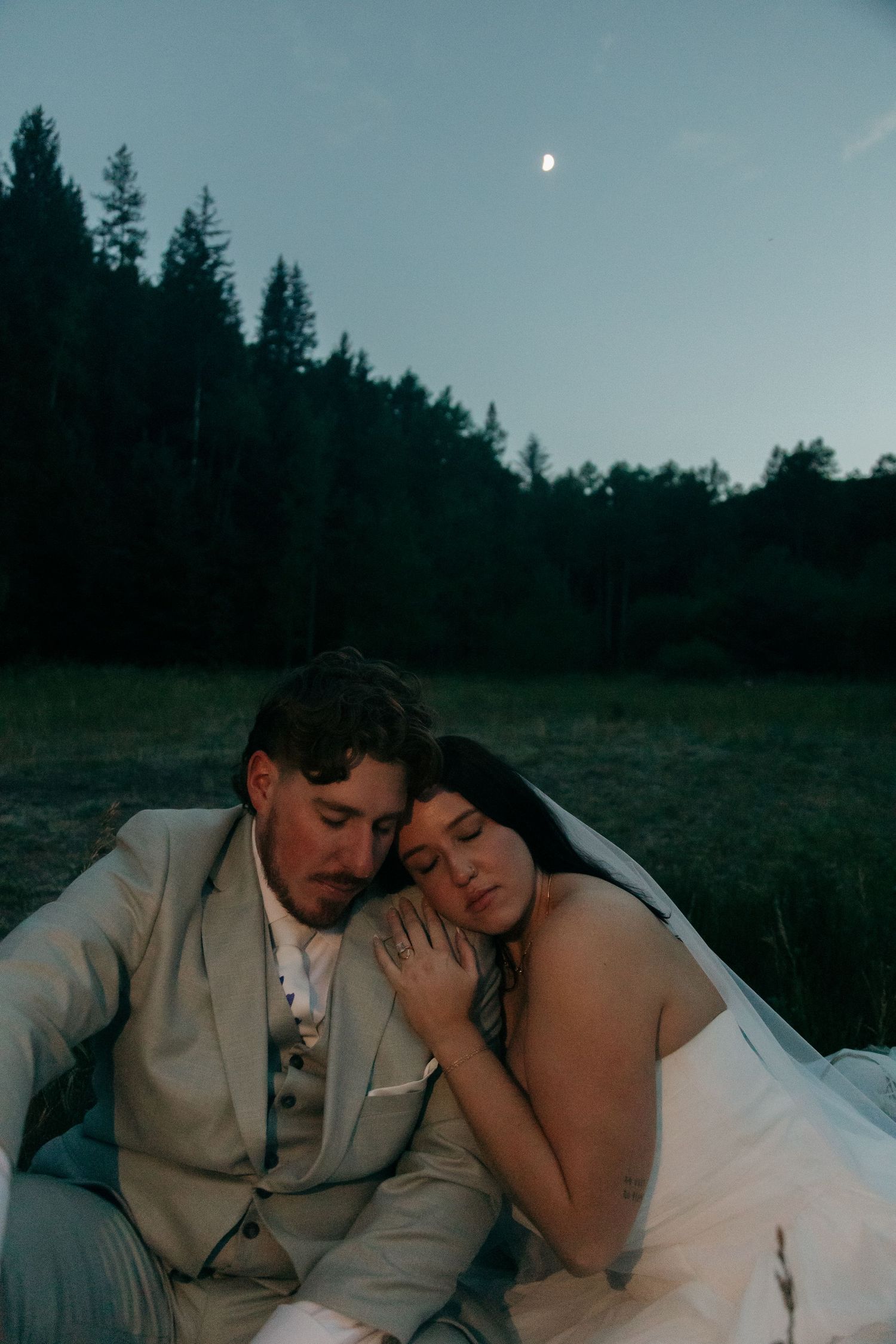 Bride rests her head on groom's shoulder as they sit together in a moonlit meadow during their romantic outdoor elopement.