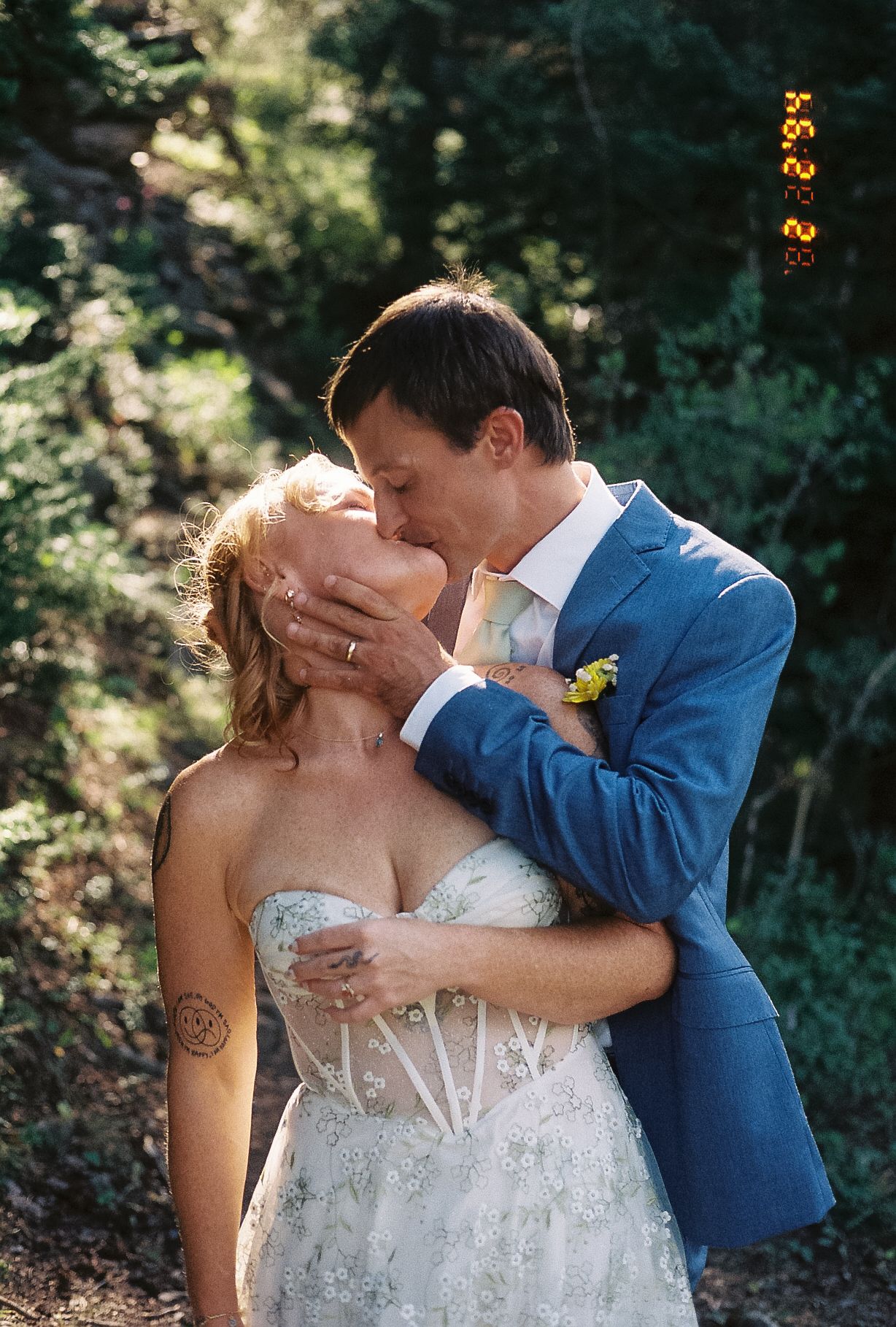Groom in blue suit kisses tattooed bride in strapless lace gown outdoors with lush green trees behind them.