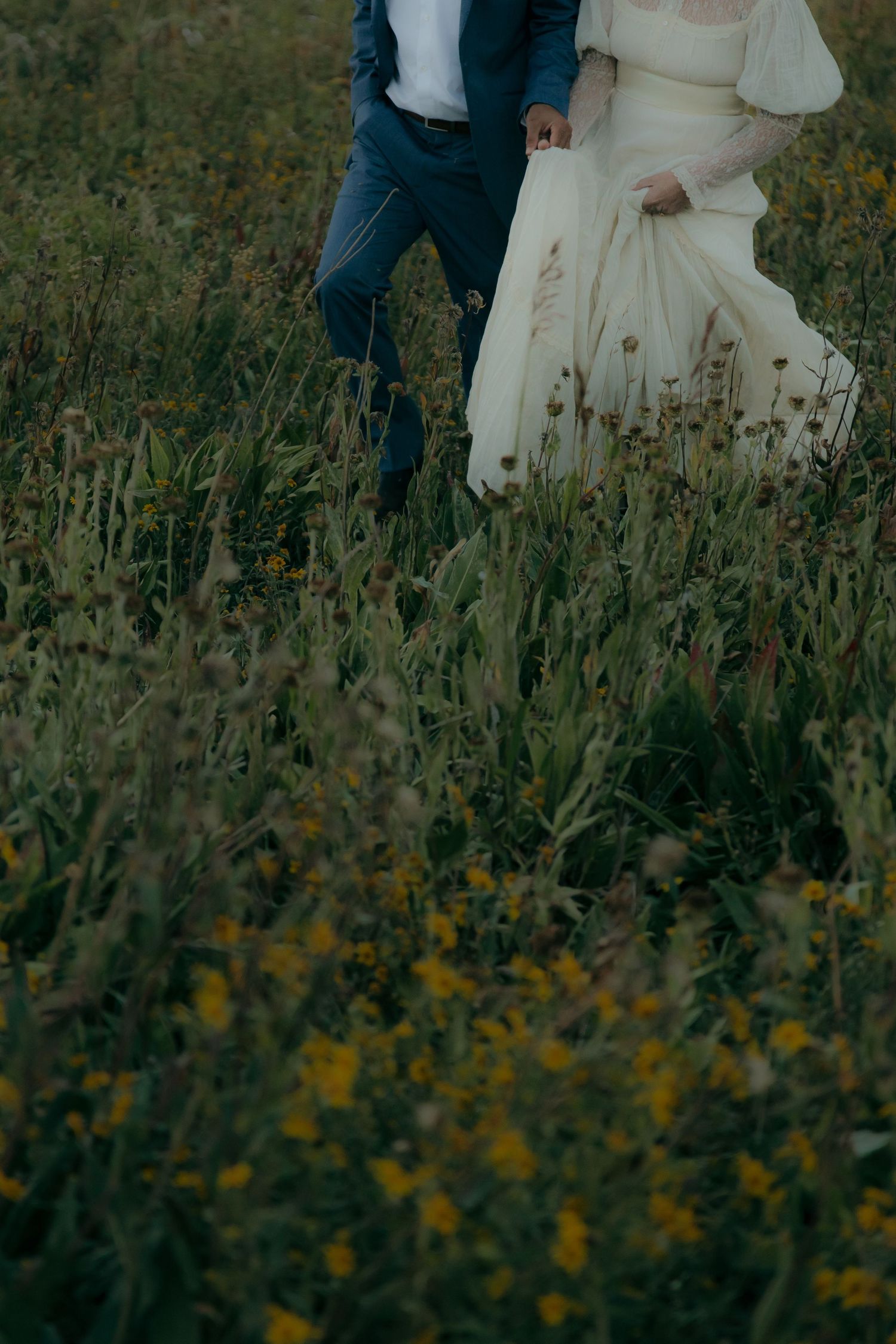 A bride in a white gown and groom in a blue suit walk hand in hand through a wildflower meadow.