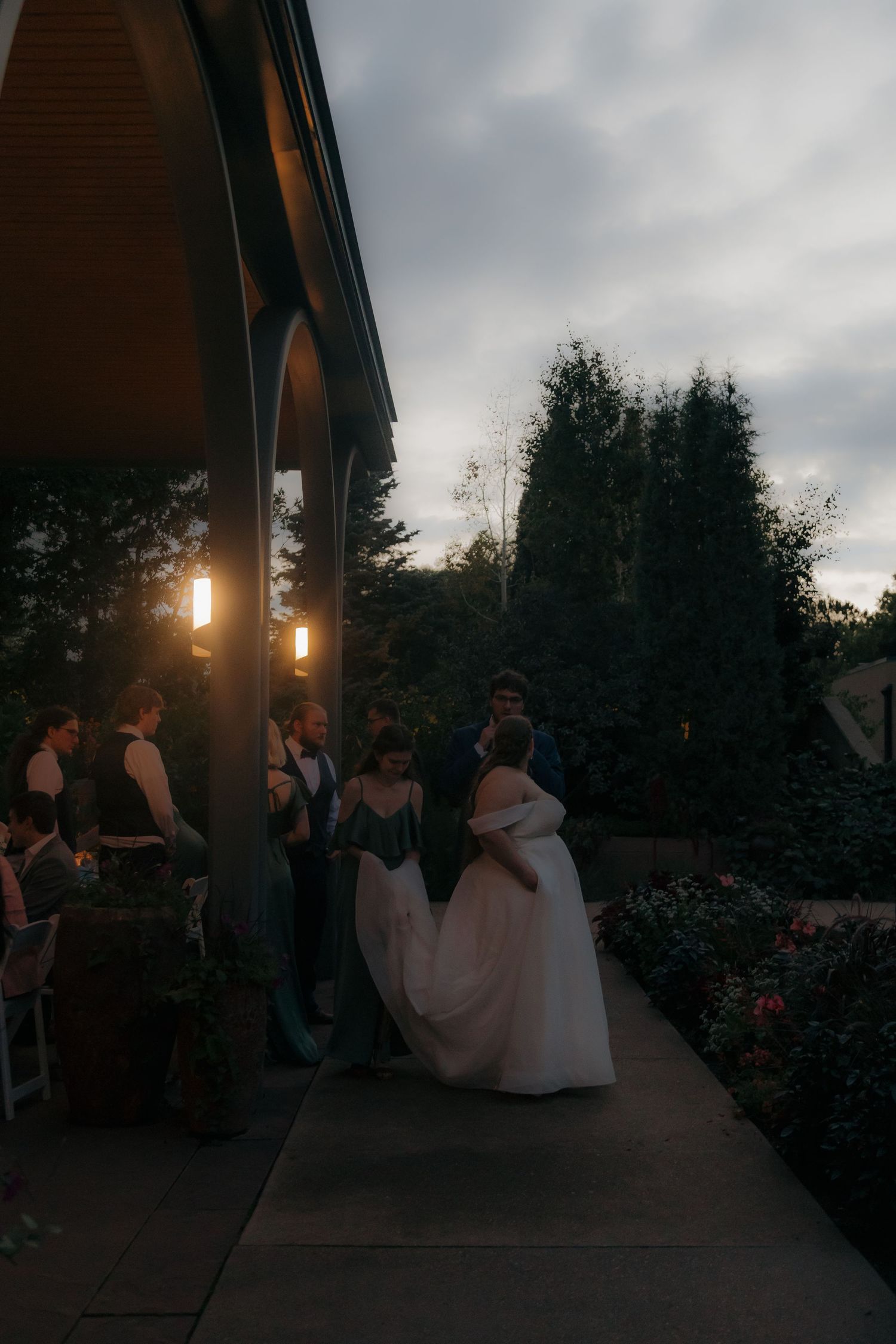 Bride in white gown walks with wedding party toward lit venue entrance at dusk.