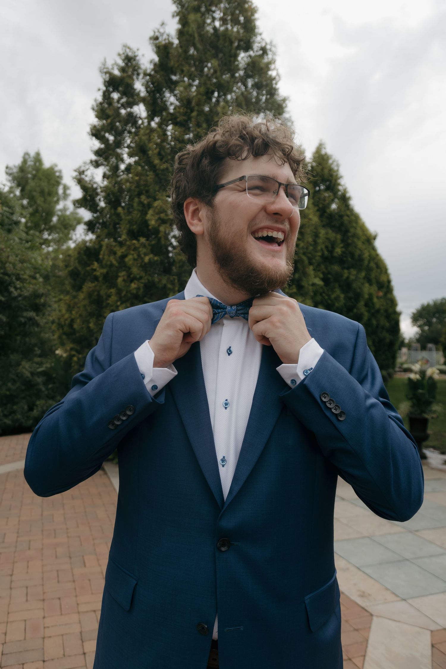 Smiling groom in blue suit adjusting blue bow tie outdoors with trees in background.