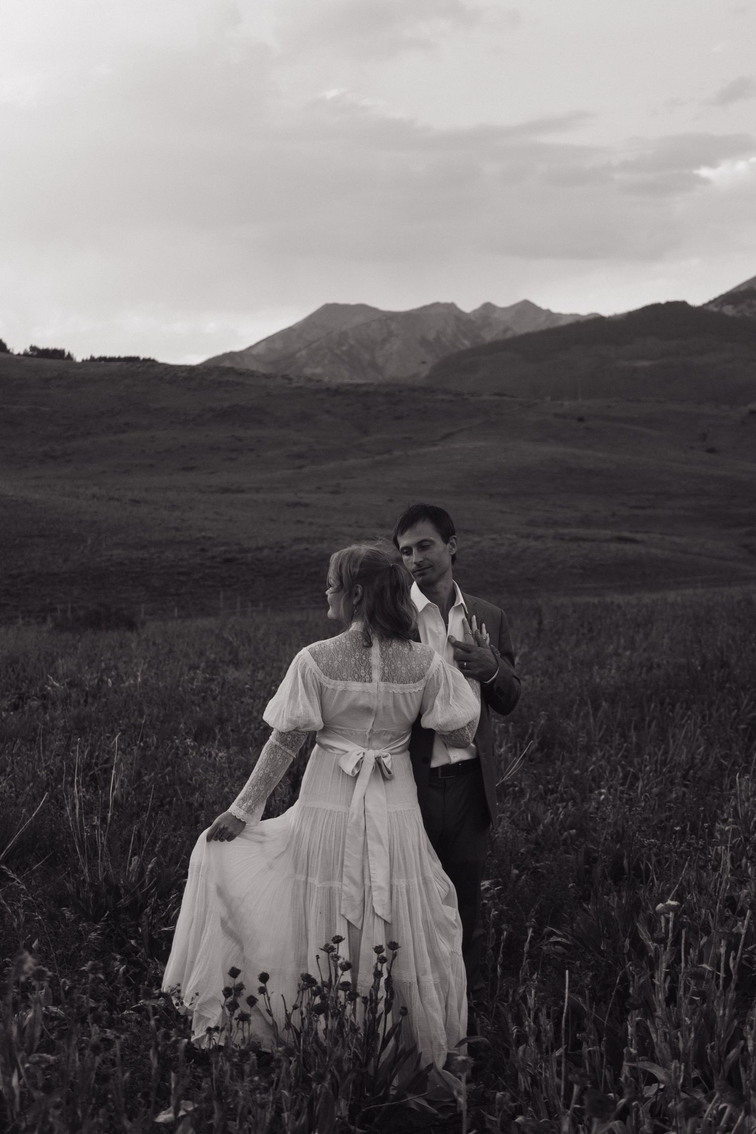 Black and white photo of bride and groom walking through a mountain meadow with dramatic peaks in the background.