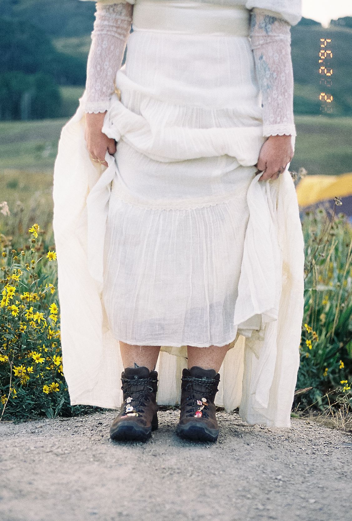 Bride in white lace gown lifting skirt to reveal black embroidered hiking boots among yellow wildflowers.