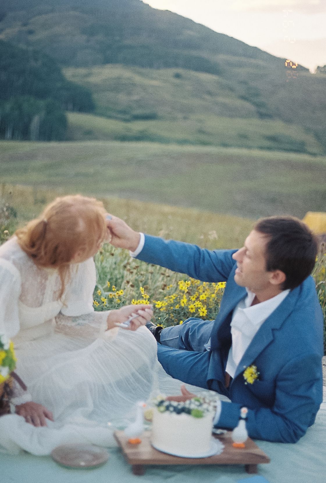 Groom in blue suit tenderly touches bride's red hair during outdoor mountain meadow wedding picnic.