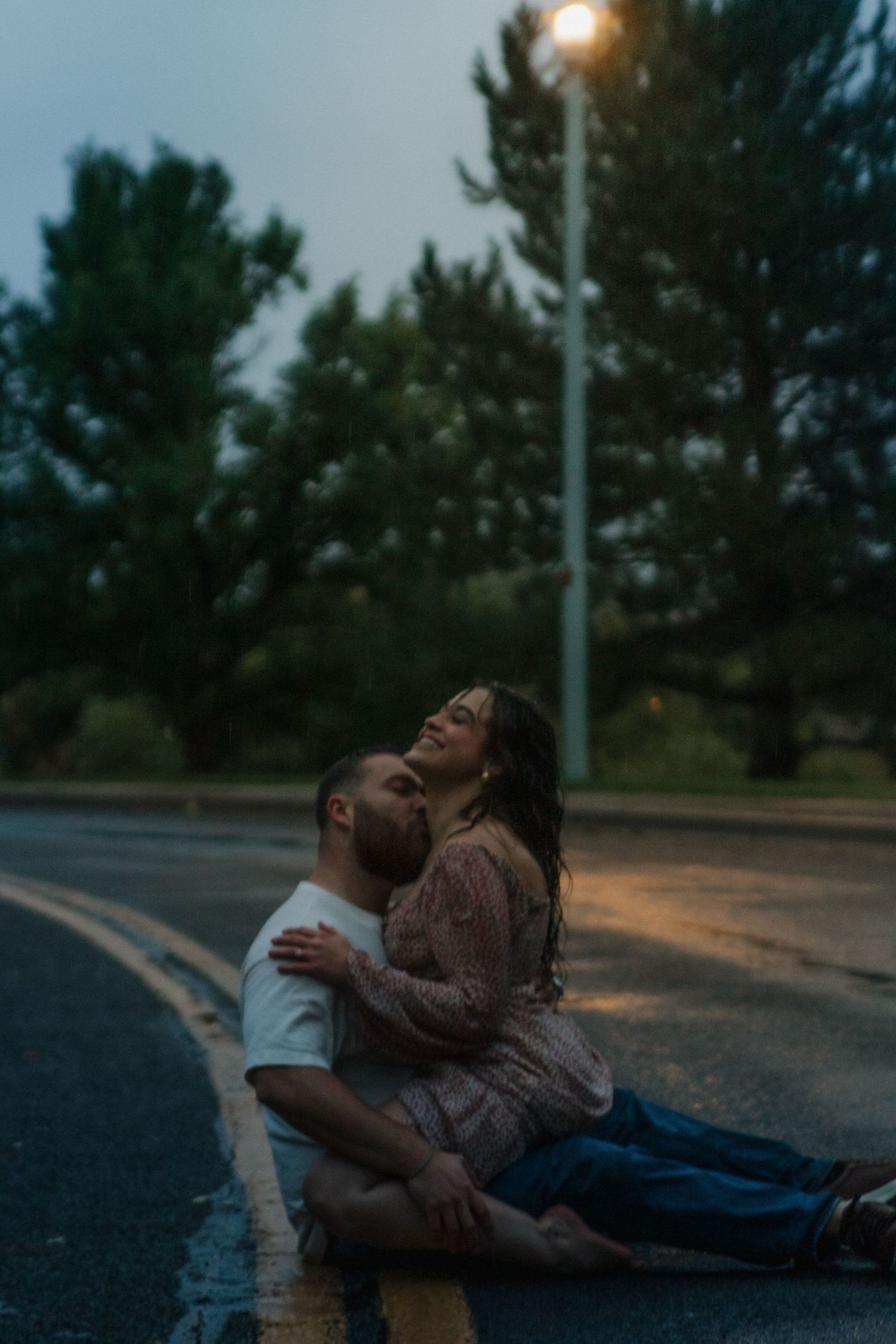A couple sits intimately on a wet road at dusk under a glowing streetlight with pine trees in the background.
