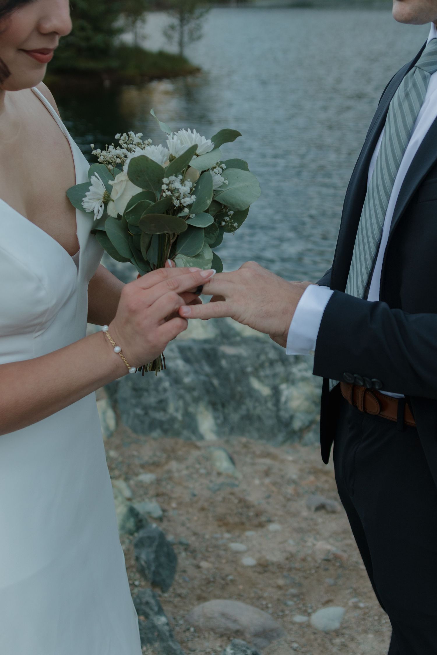 A wedding couple stands by a lake exchanging white flowers during an intimate lakeside ceremony.