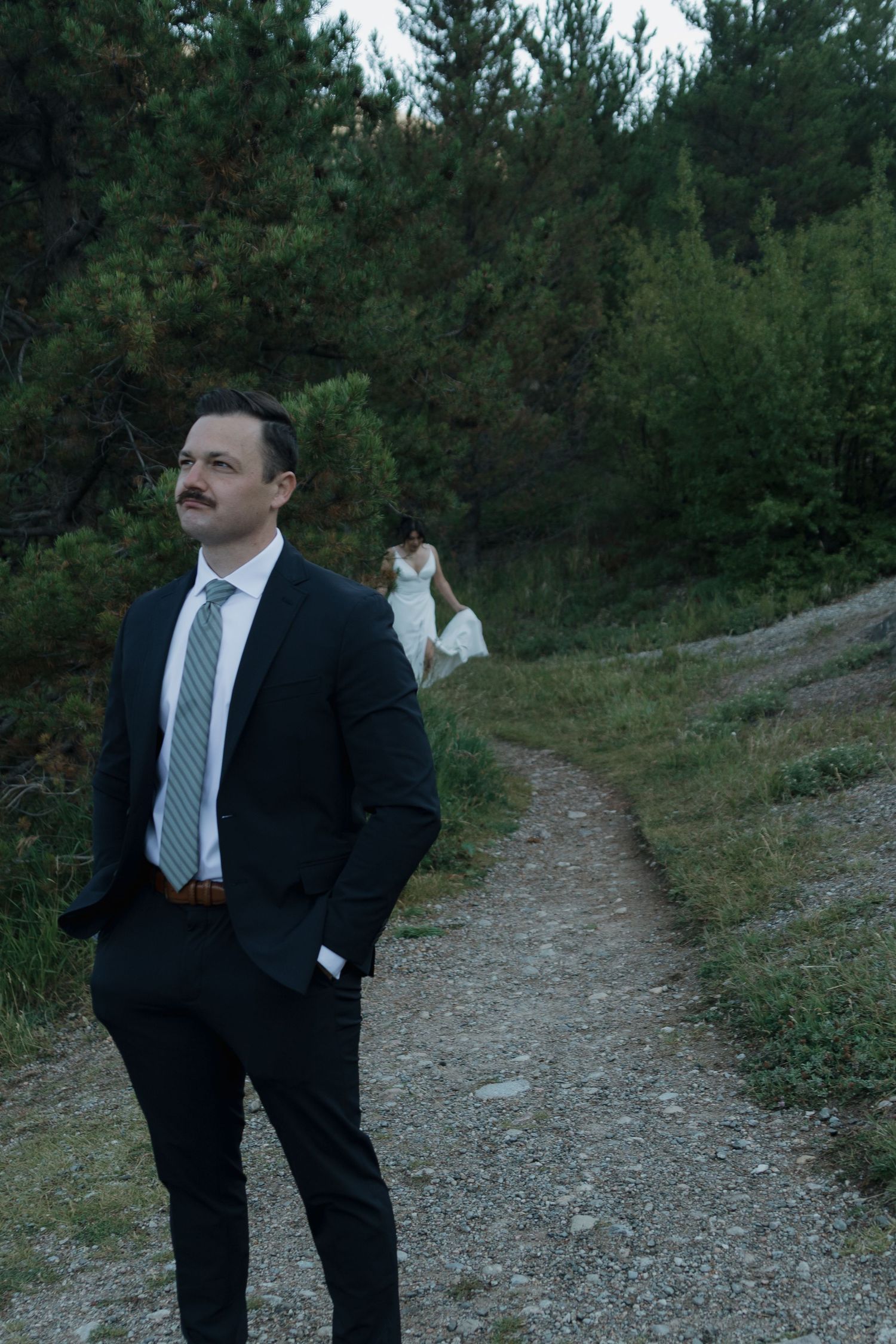 A couple in formal attire walks down a gravel path surrounded by green trees and foliage.