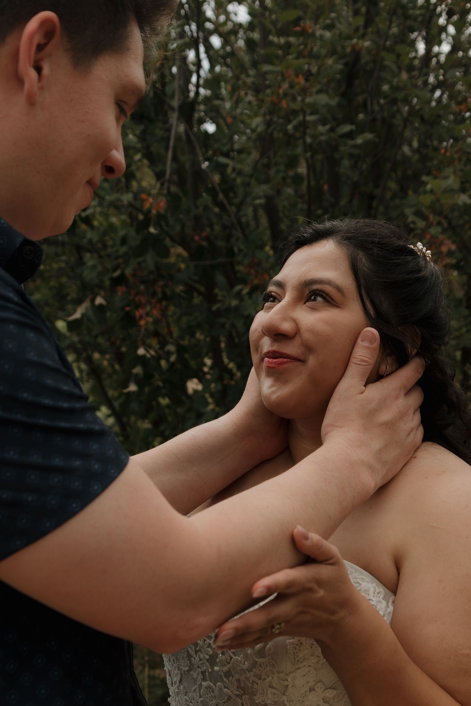 Close-up romantic wedding couple moment with hands gently caressing faces in natural outdoor setting.