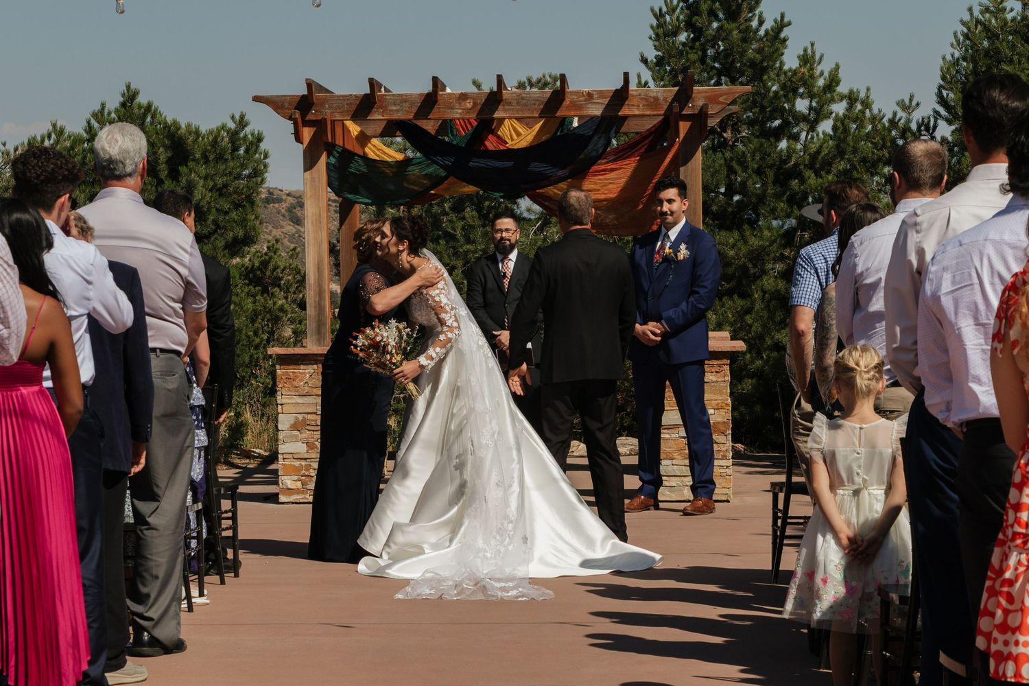 Wedding ceremony under wooden pergola with mountain views and gathered guests.