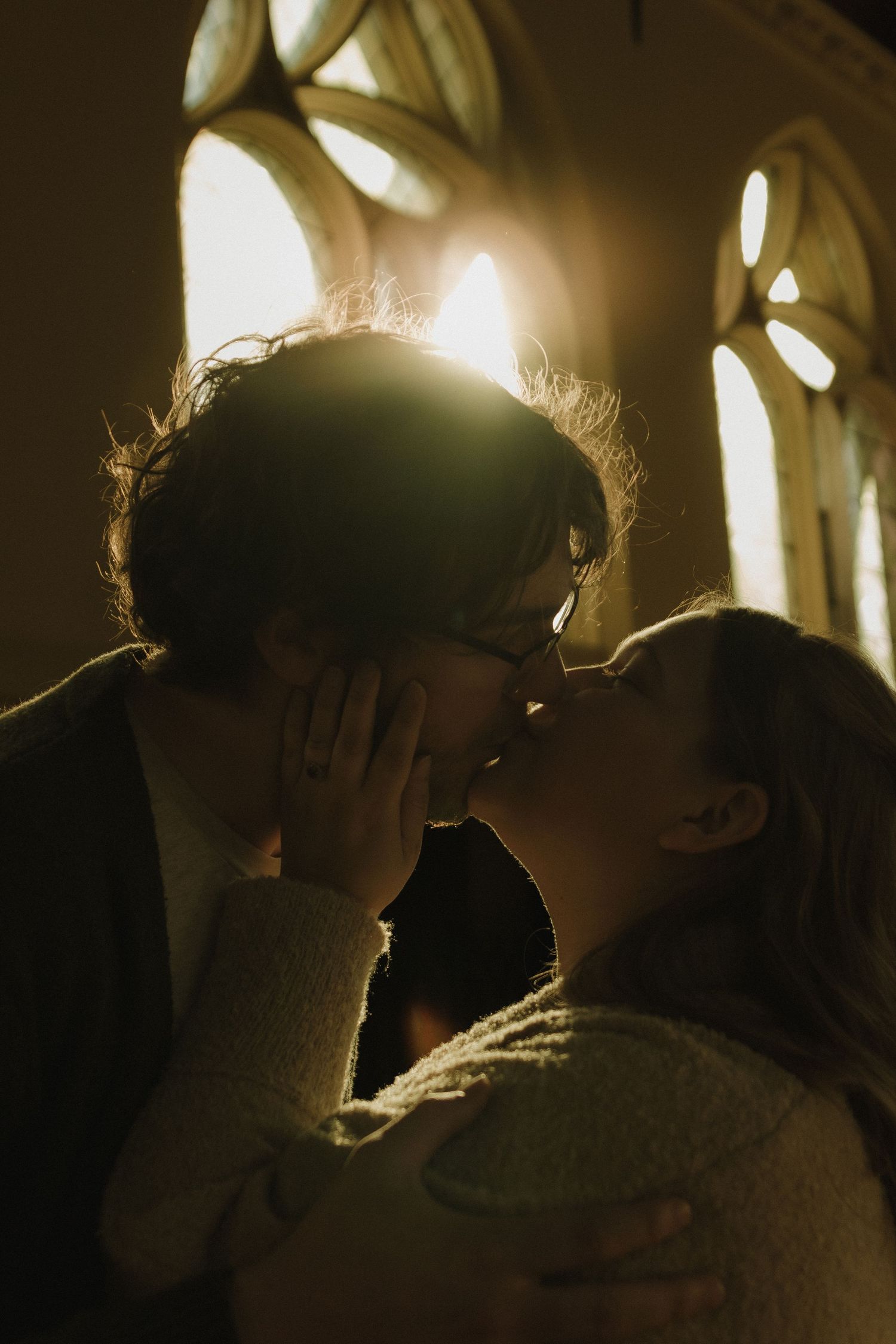 Couple sharing a romantic kiss backlit by golden sunlight streaming through gothic church windows.