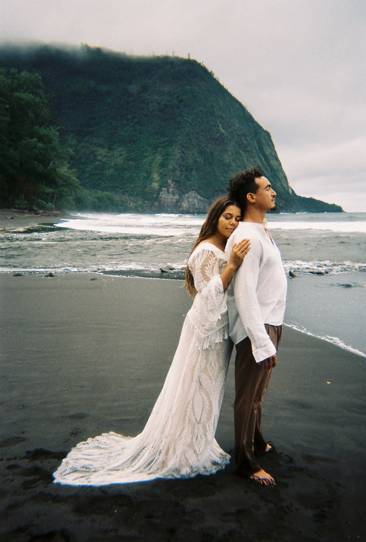 Bride in lace wedding gown embracing groom from behind on black sand beach with misty green cliffs.