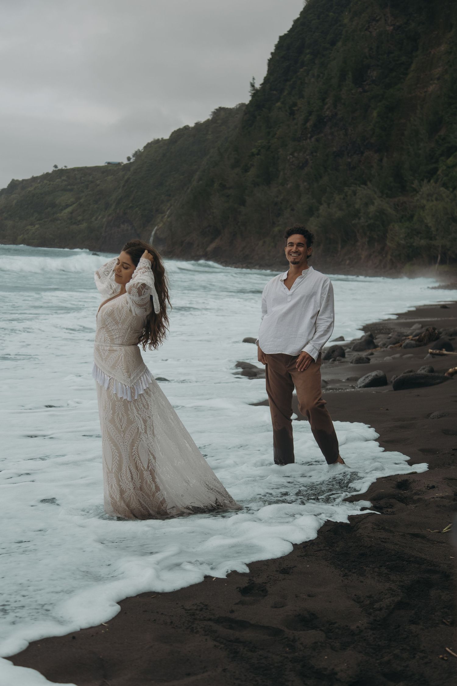 Couple standing in ocean waves on black sand beach, woman in white lace dress and man in white shirt smiling.