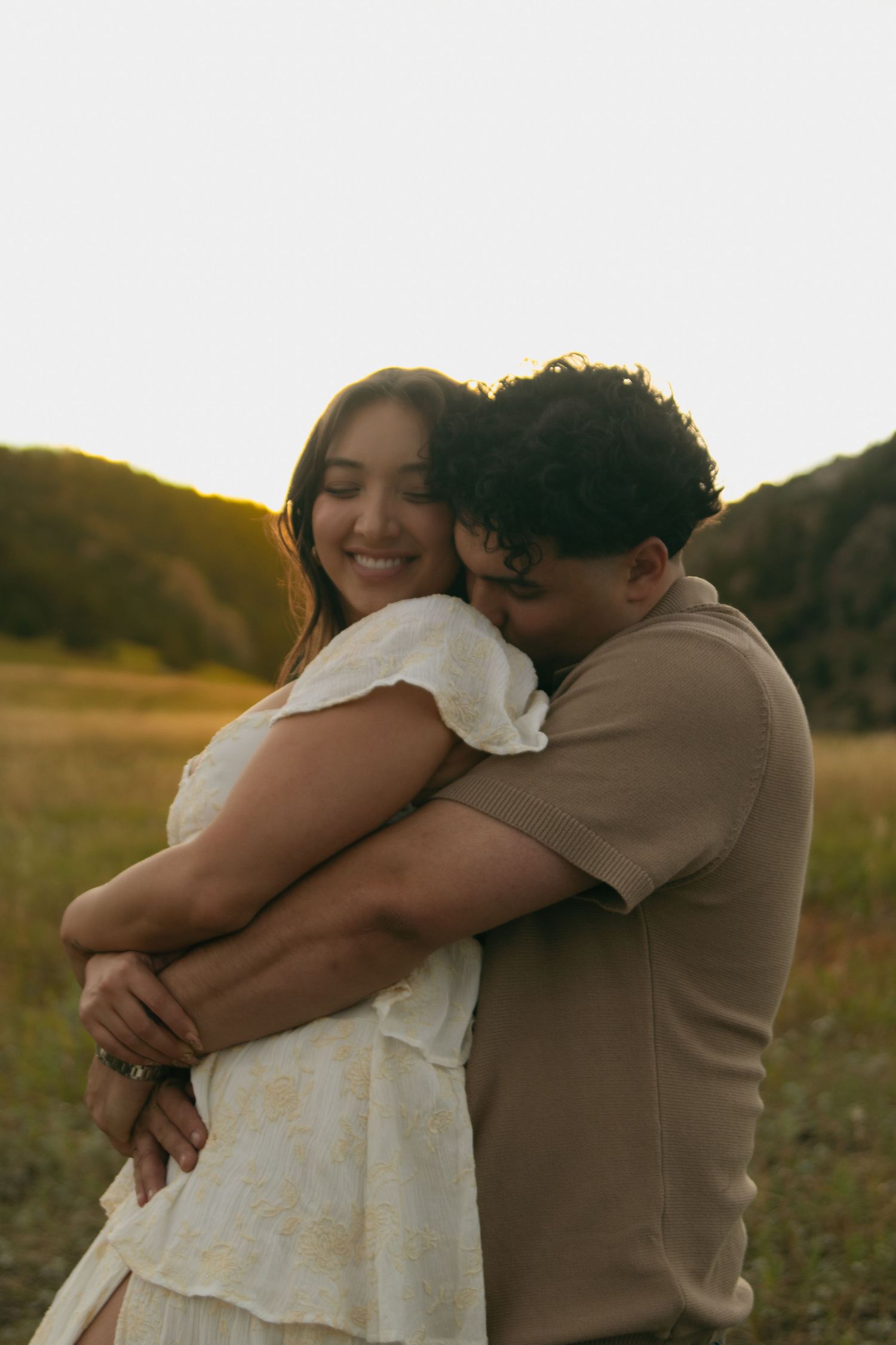 A smiling woman in a white dress embraced from behind by her partner during a sunset engagement photo.