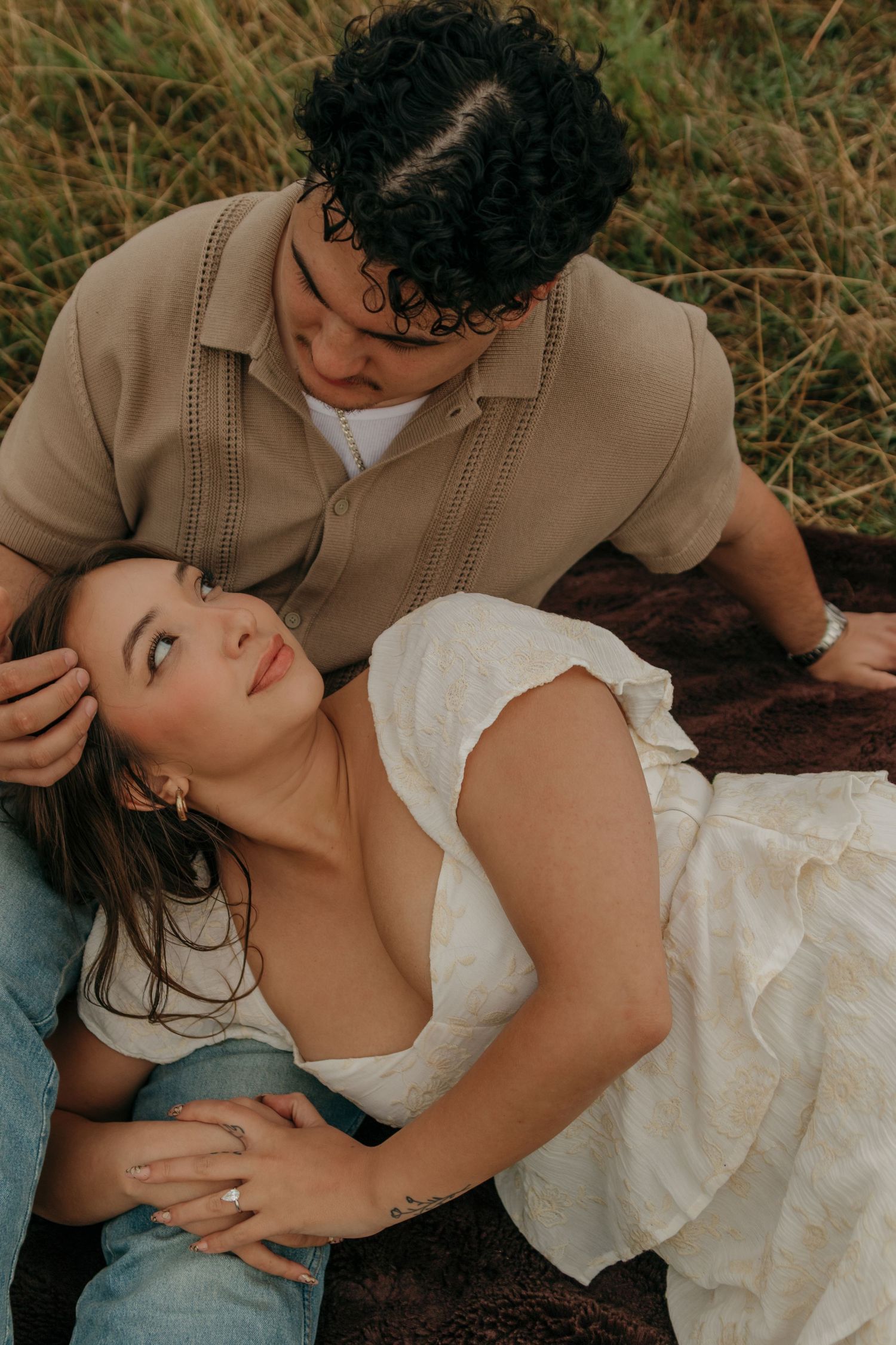 Couple relaxes on a blanket in a grassy field, woman gazing up at her partner tenderly.