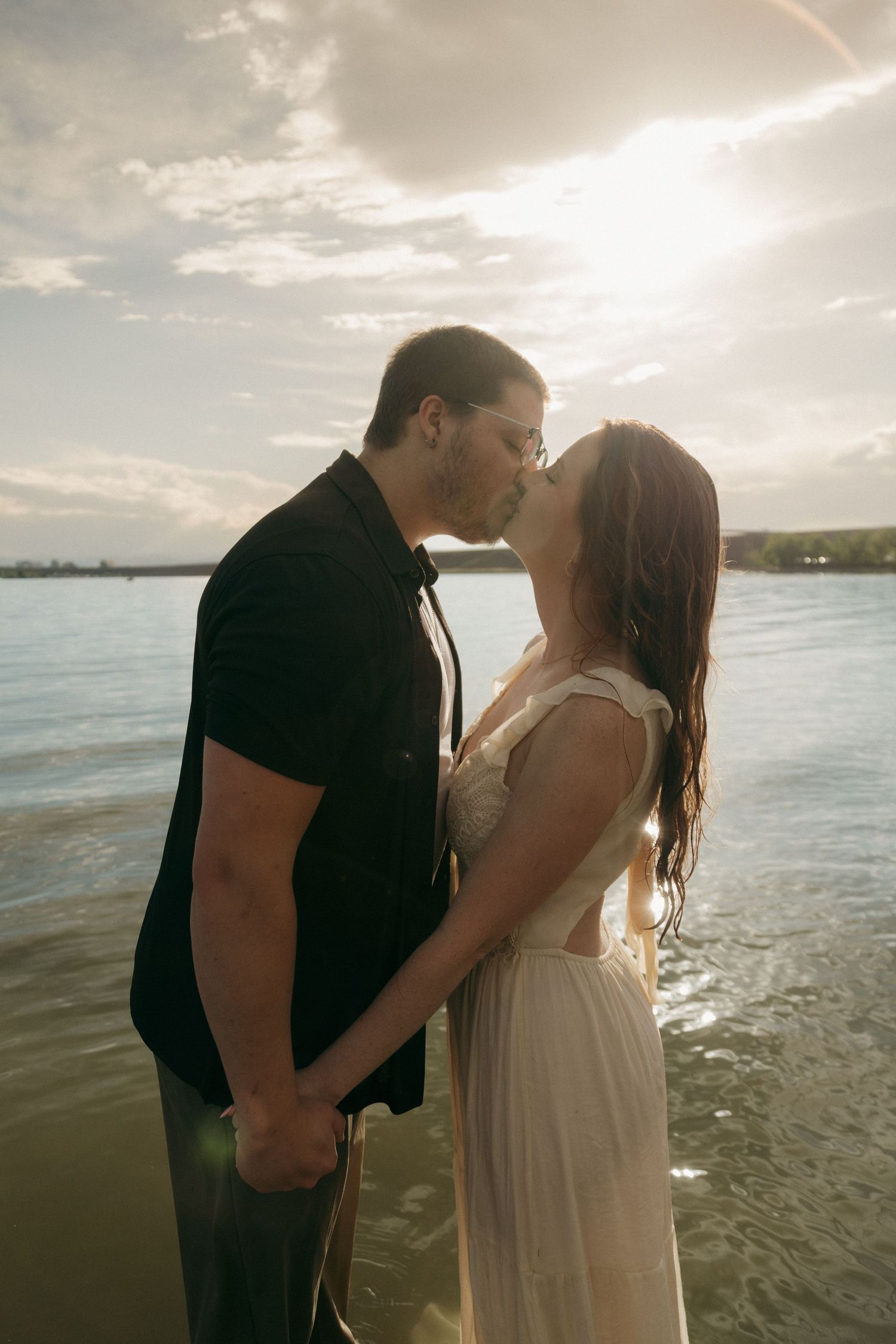 Couple sharing a kiss while standing in lake water at sunset, woman in white bridal dress.