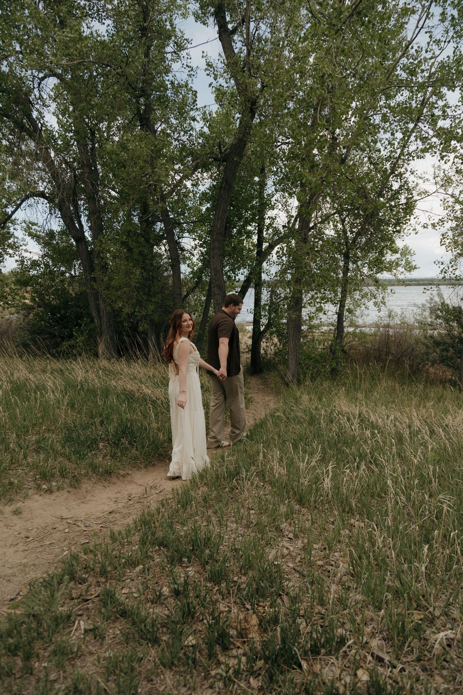 Couple holding hands on a sandy path surrounded by lush green trees near a lake during an outdoor engagement session.