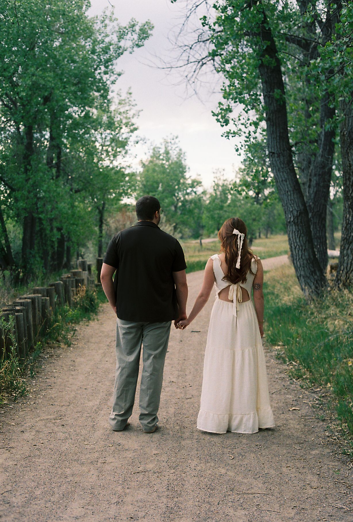 Couple holding hands walking down a tree-lined dirt path, woman in white open-back dress.