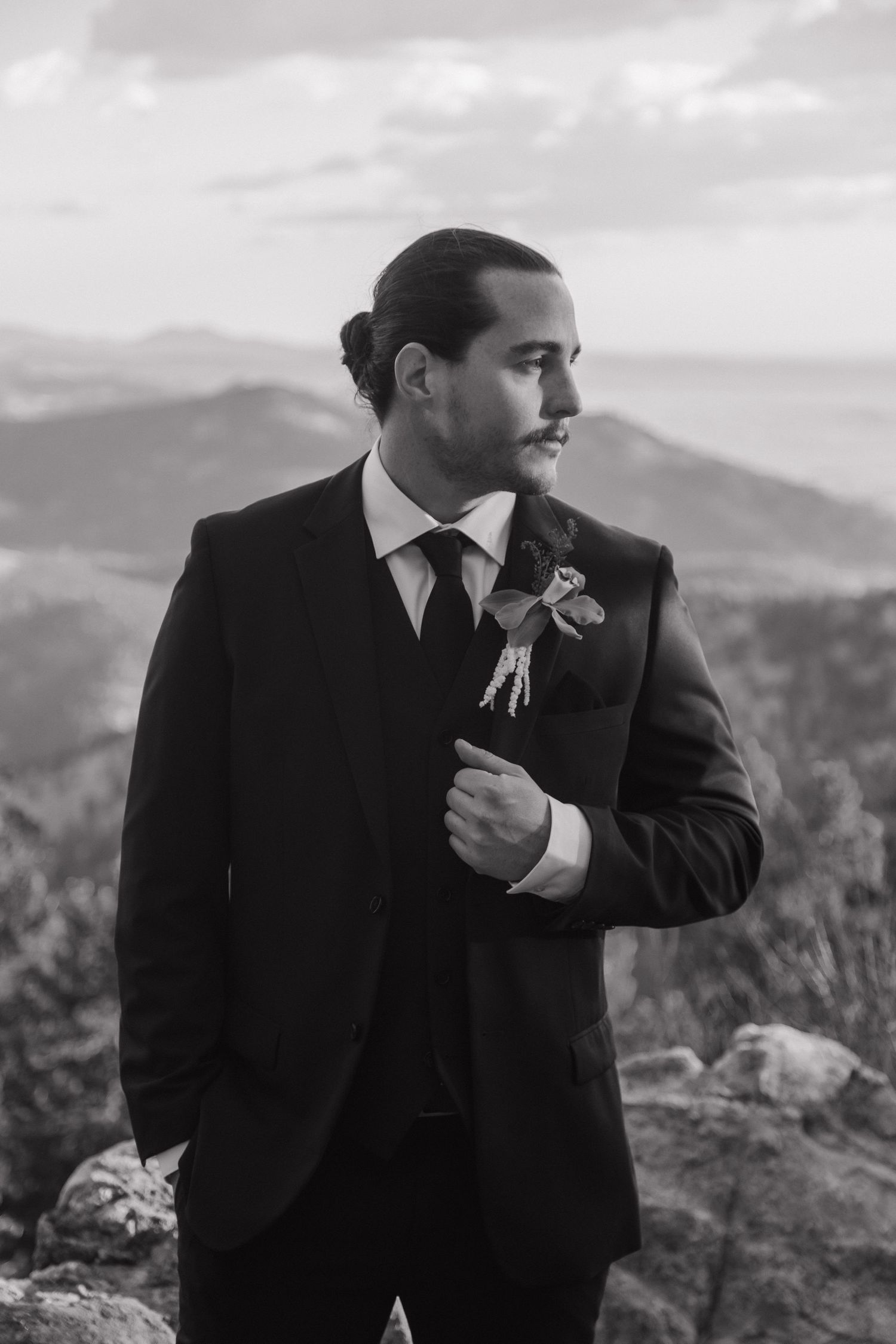 Dapper groom in black suit with boutonniere poses against scenic mountain backdrop in black and white.