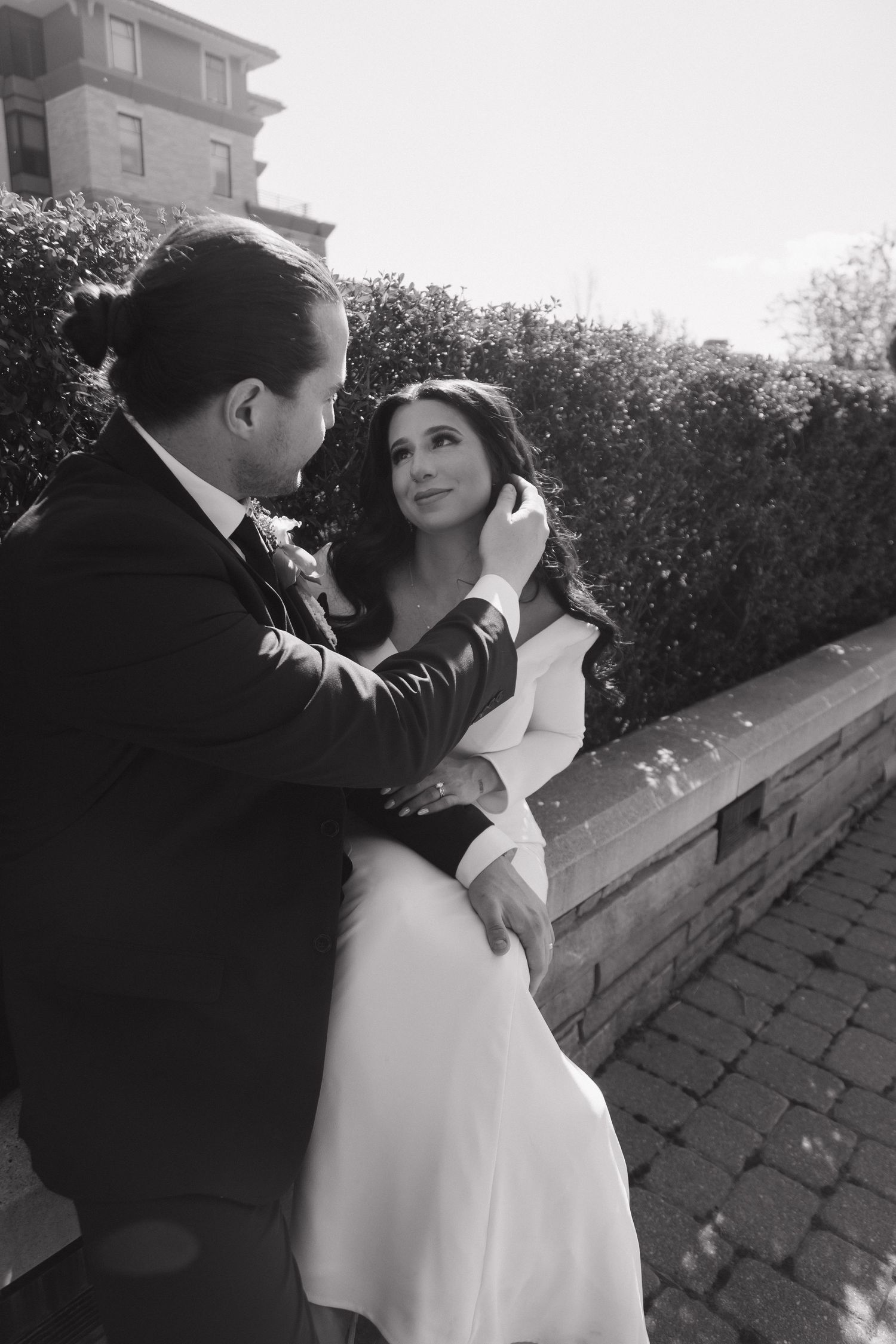 Black and white photo of groom tenderly touching brides face as she gazes up at him on a garden wall.