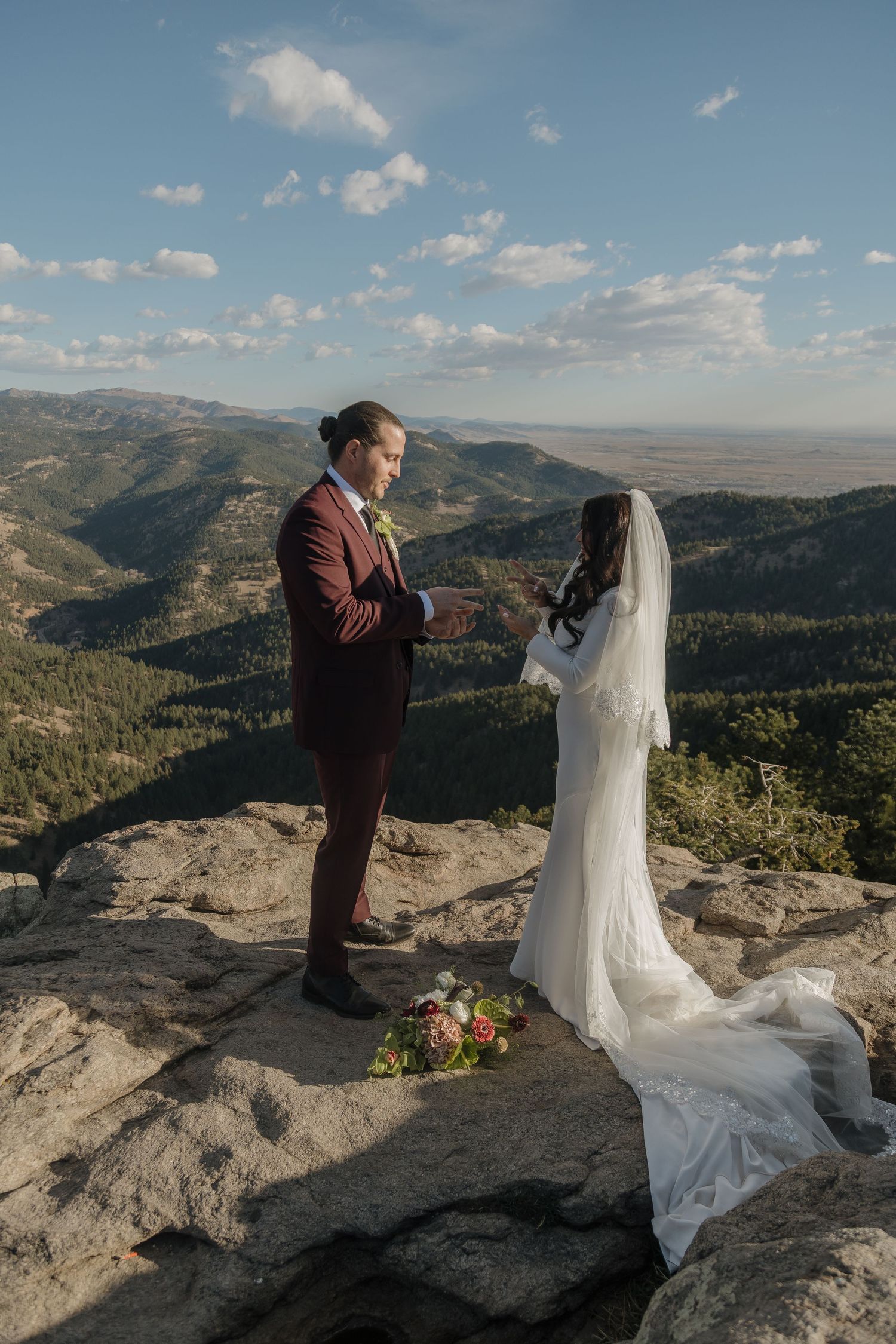 Groom in burgundy suit reads vows to bride in white gown on rocky mountain overlook.