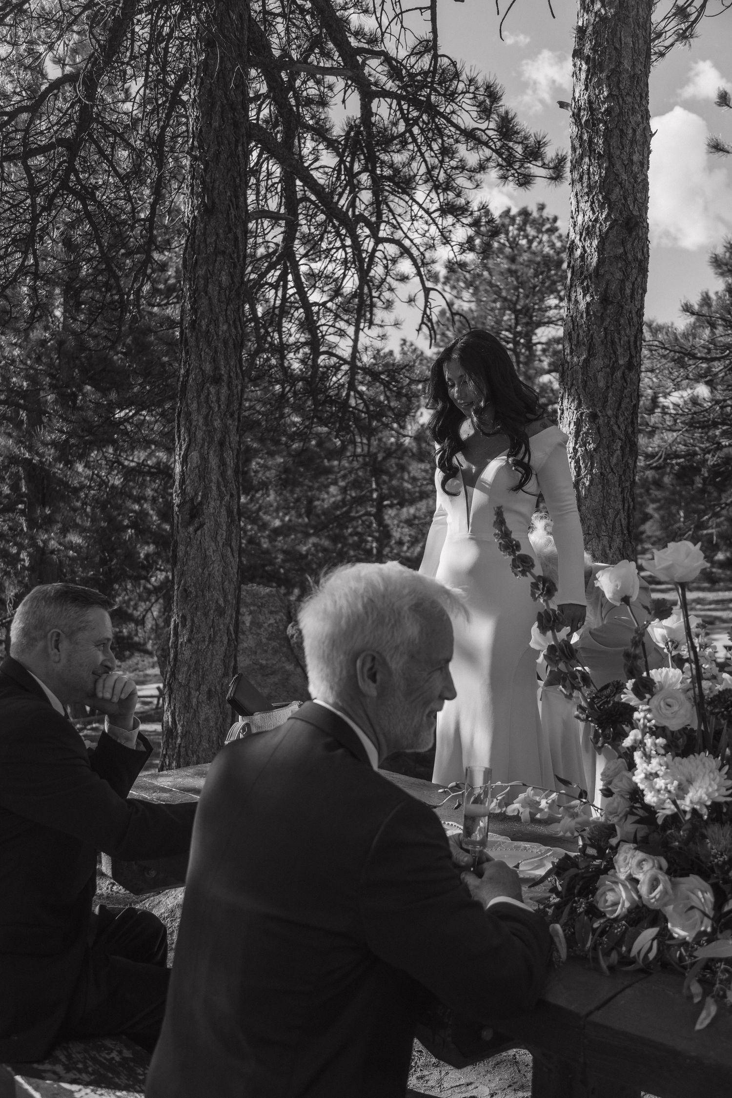 Black and white photo of bride in white gown standing at outdoor wedding table surrounded by guests and flowers.