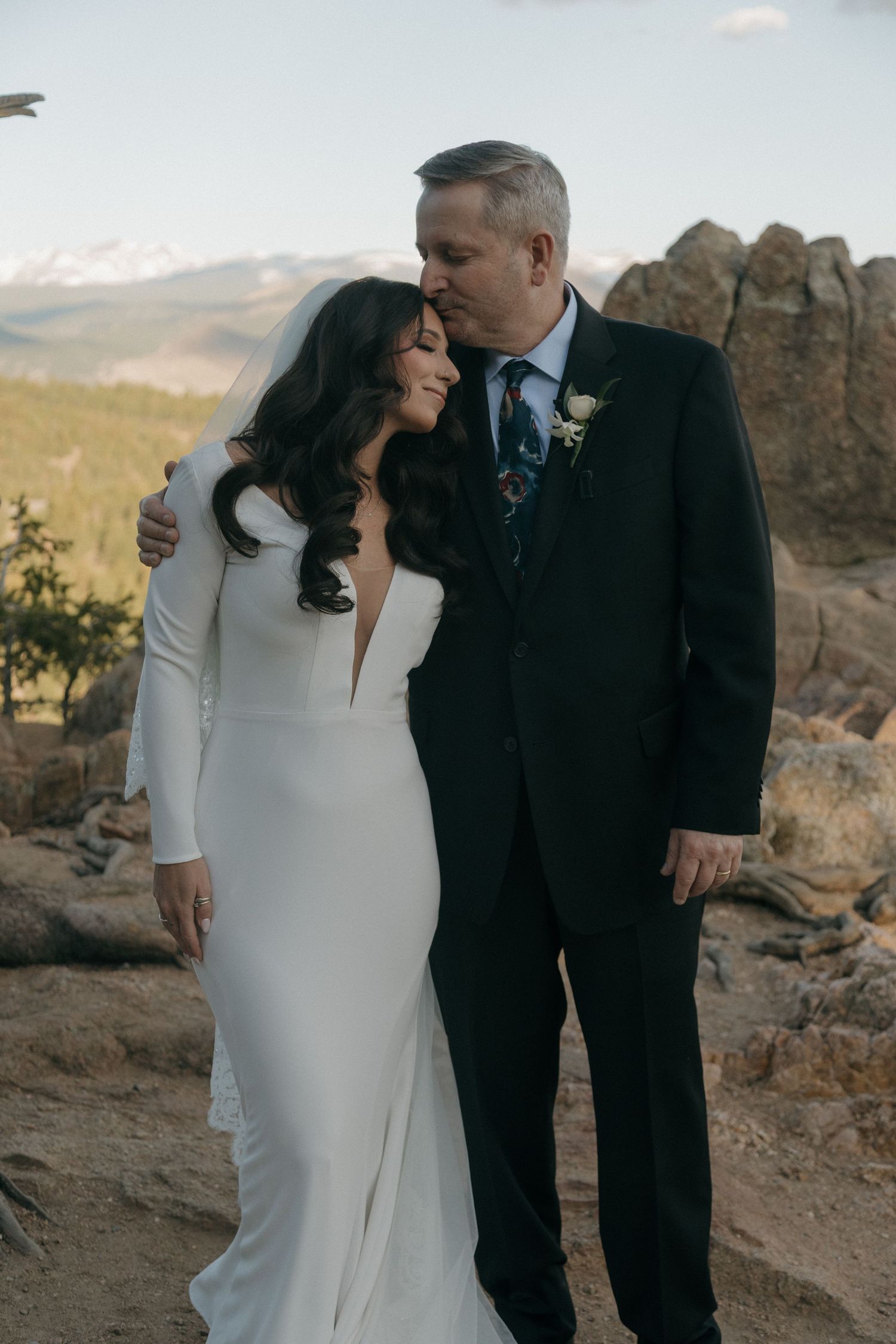 Bride in white fitted gown and groom in dark suit share tender moment at rocky mountain wedding venue.