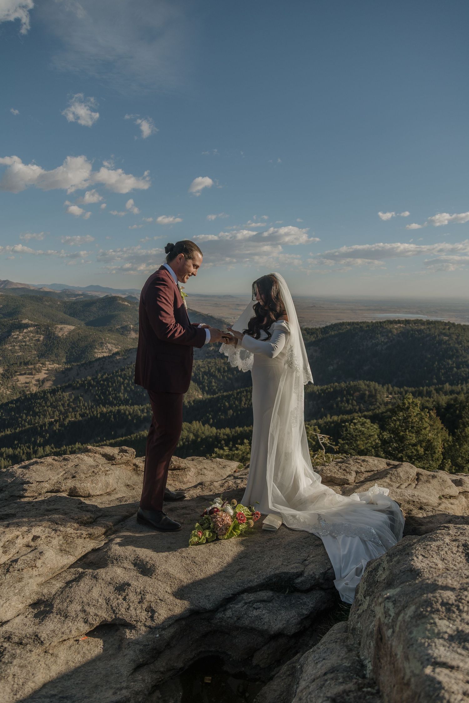 Bride and groom hold hands during vow exchange on scenic mountain summit with sweeping Colorado landscape below.
