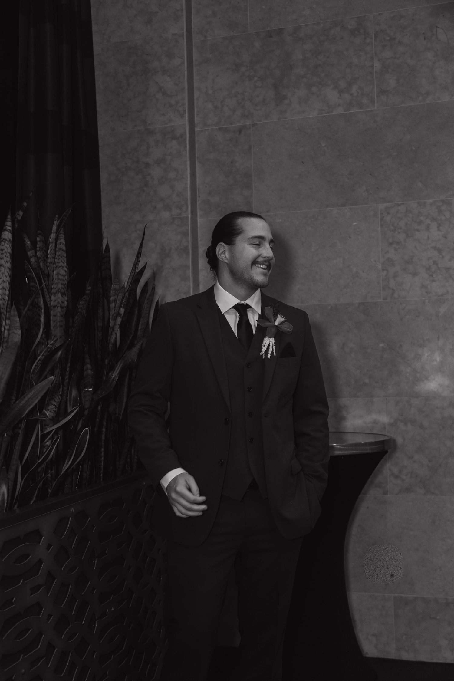 Smiling groom in black three-piece suit with boutonniere poses against stone wall in black and white photo.