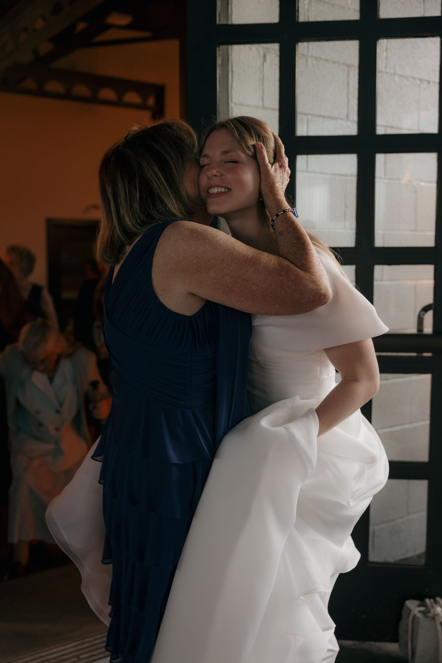 Bride in white gown shares emotional hug with guest in navy dress at wedding reception.