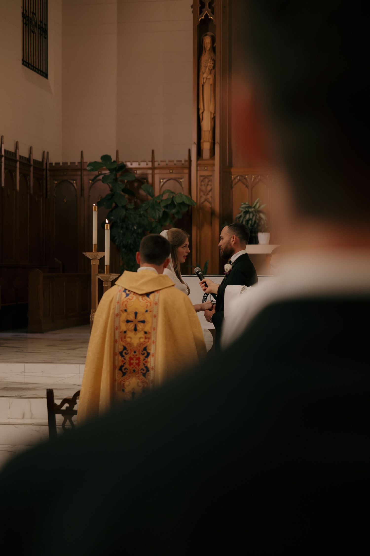 Priest in gold vestments officiates wedding ceremony as bride and groom exchange vows at altar.