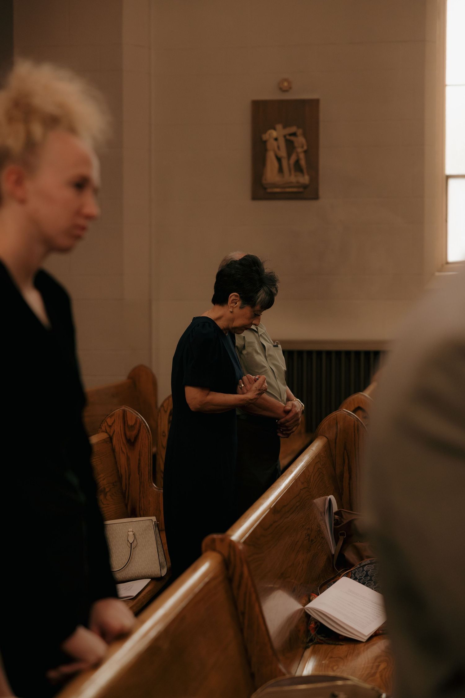 Wedding guest bows head in prayer during church ceremony beside wooden pews with religious artwork on wall.