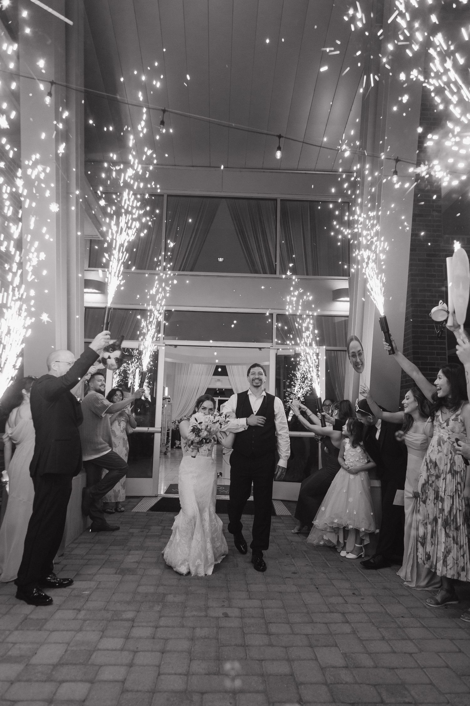 Black and white photo of bride and groom exiting wedding venue through sparkler send-off with guests.