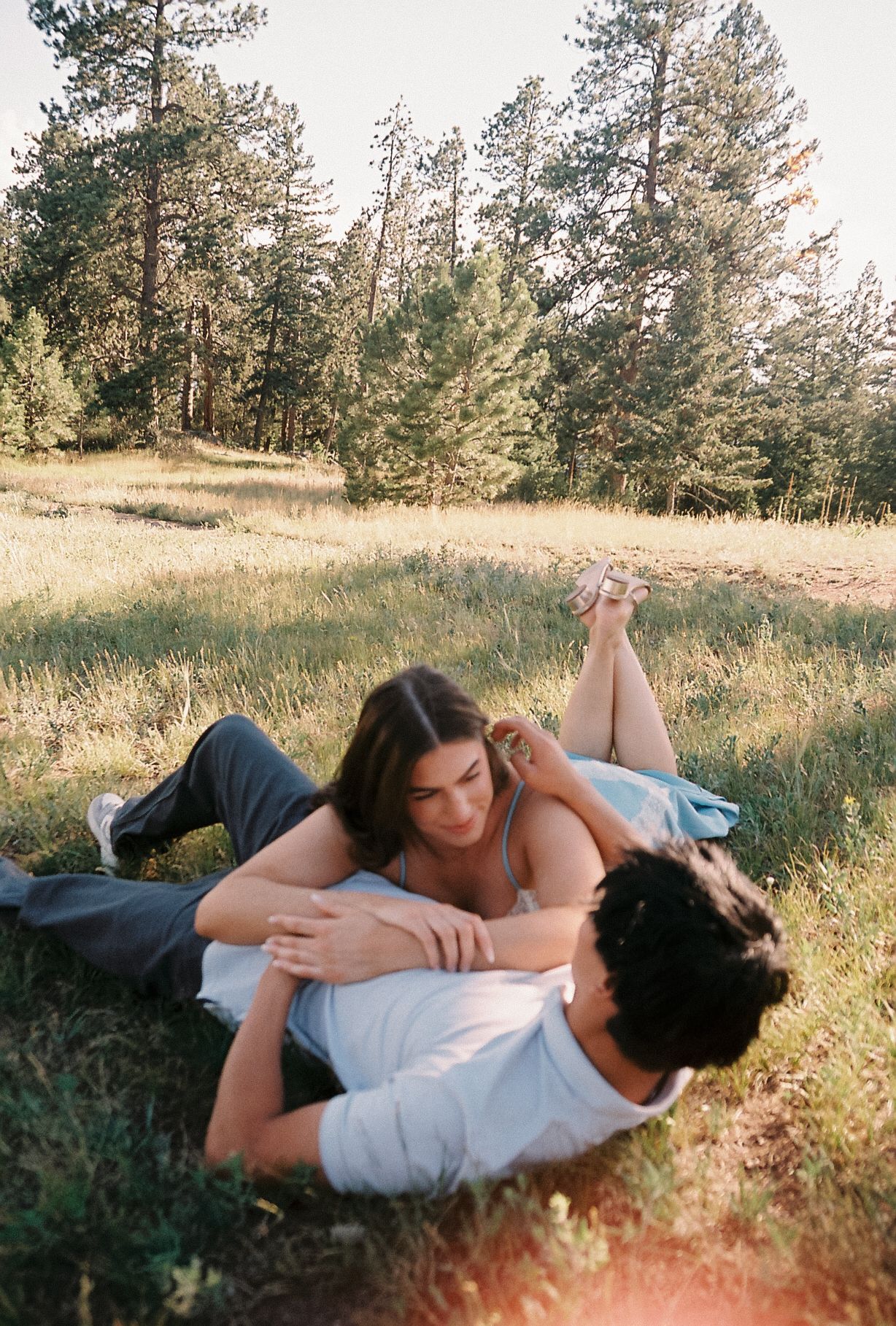 A couple lies in a sunlit meadow surrounded by pine trees, sharing a tender romantic moment in a film photography style.