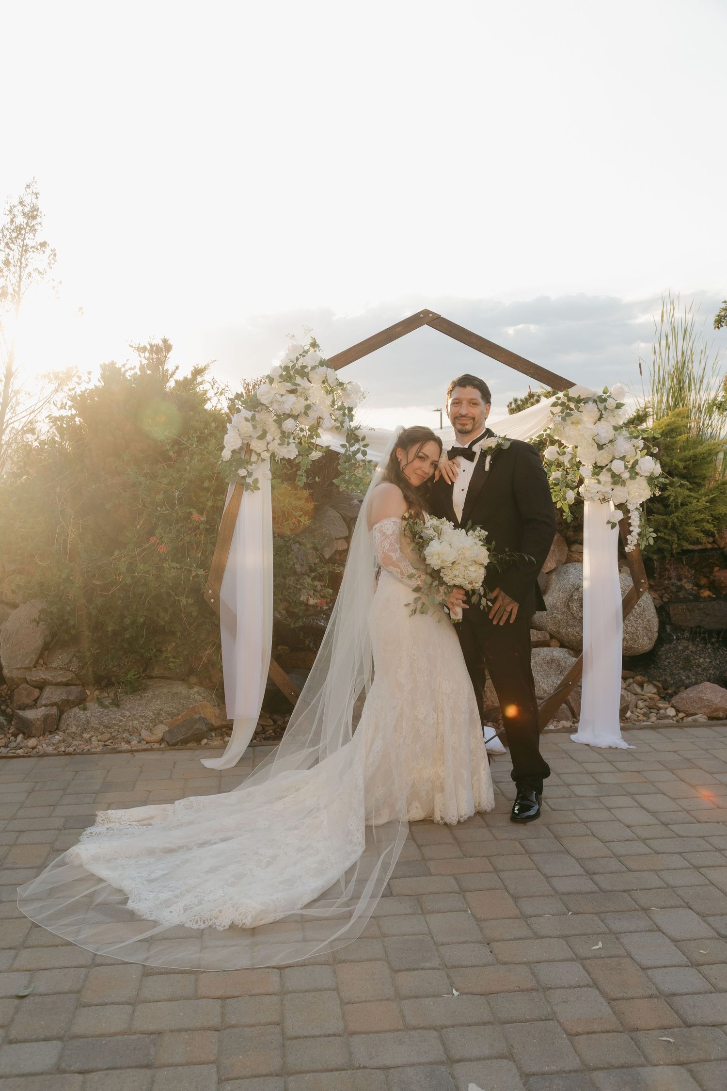 Bride and groom posing at wooden arch with white flowers and flowing fabric at sunset.