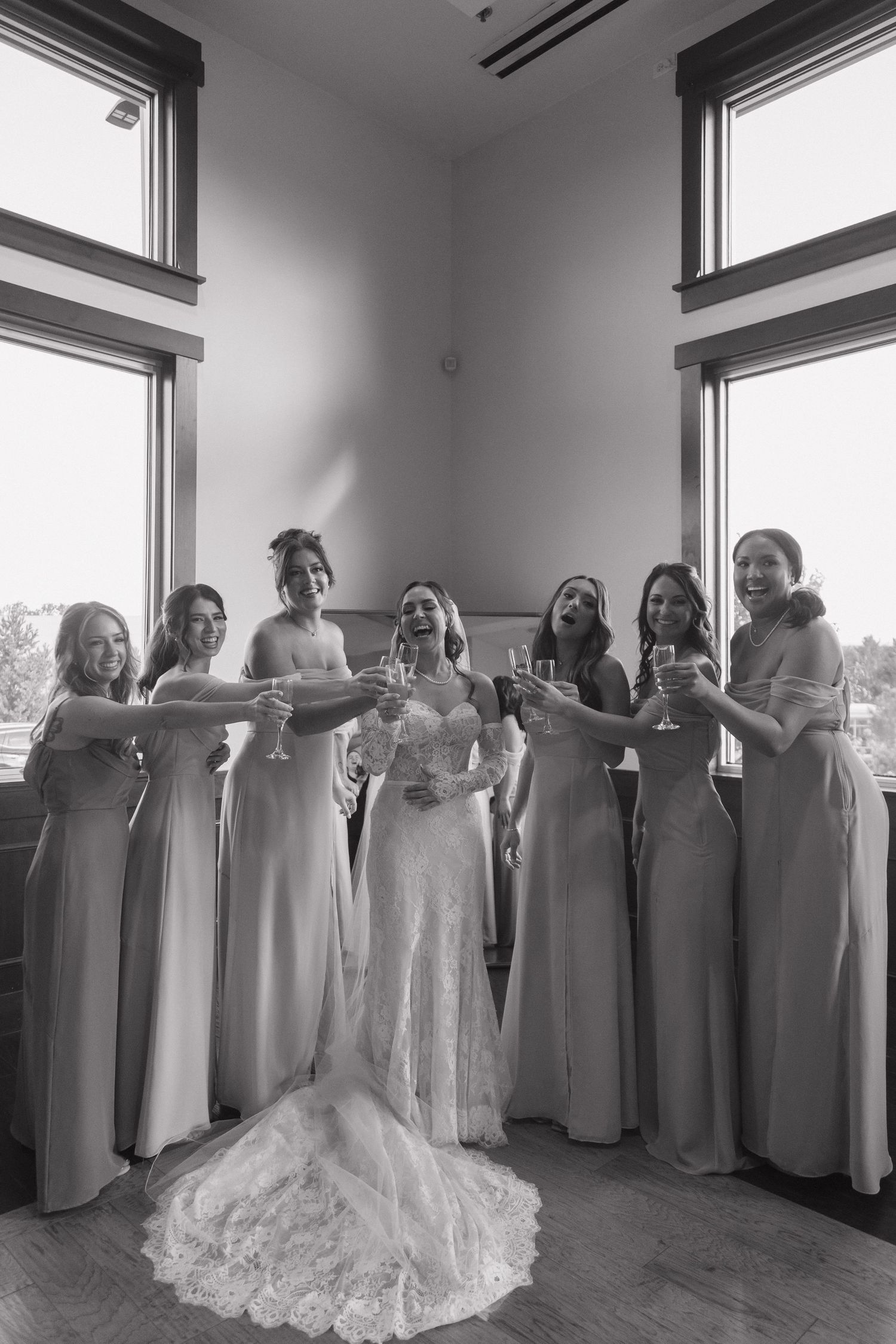 Bride and six bridesmaids in matching gowns toast with champagne glasses in black and white photo.